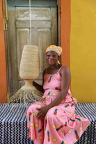 Woman in a pink dress with pineapple pattern holding a woven lampshade against a colorful background.