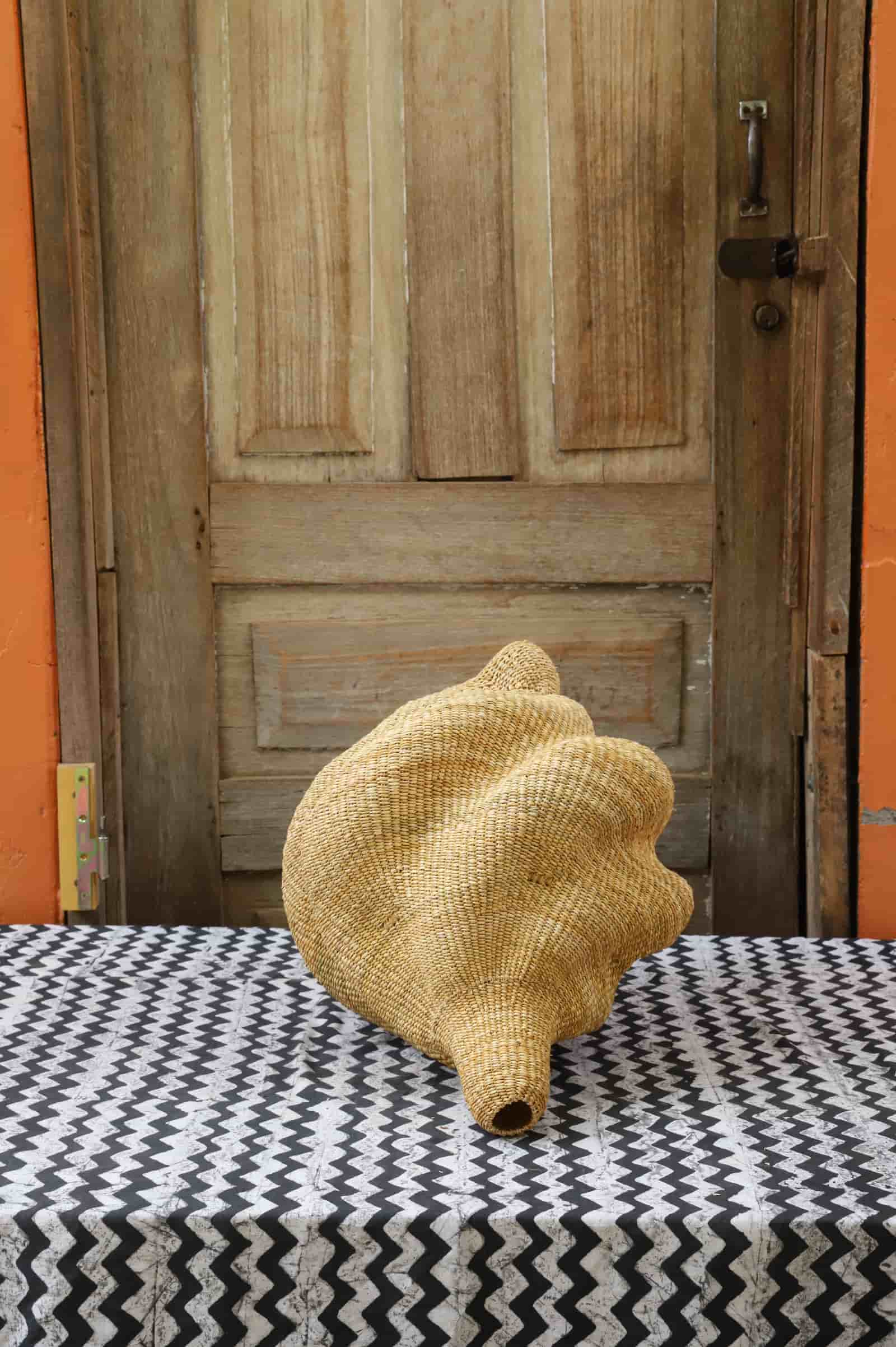 Woven conch-shaped lampshade on a black and white patterned cloth with a wooden door in the background.