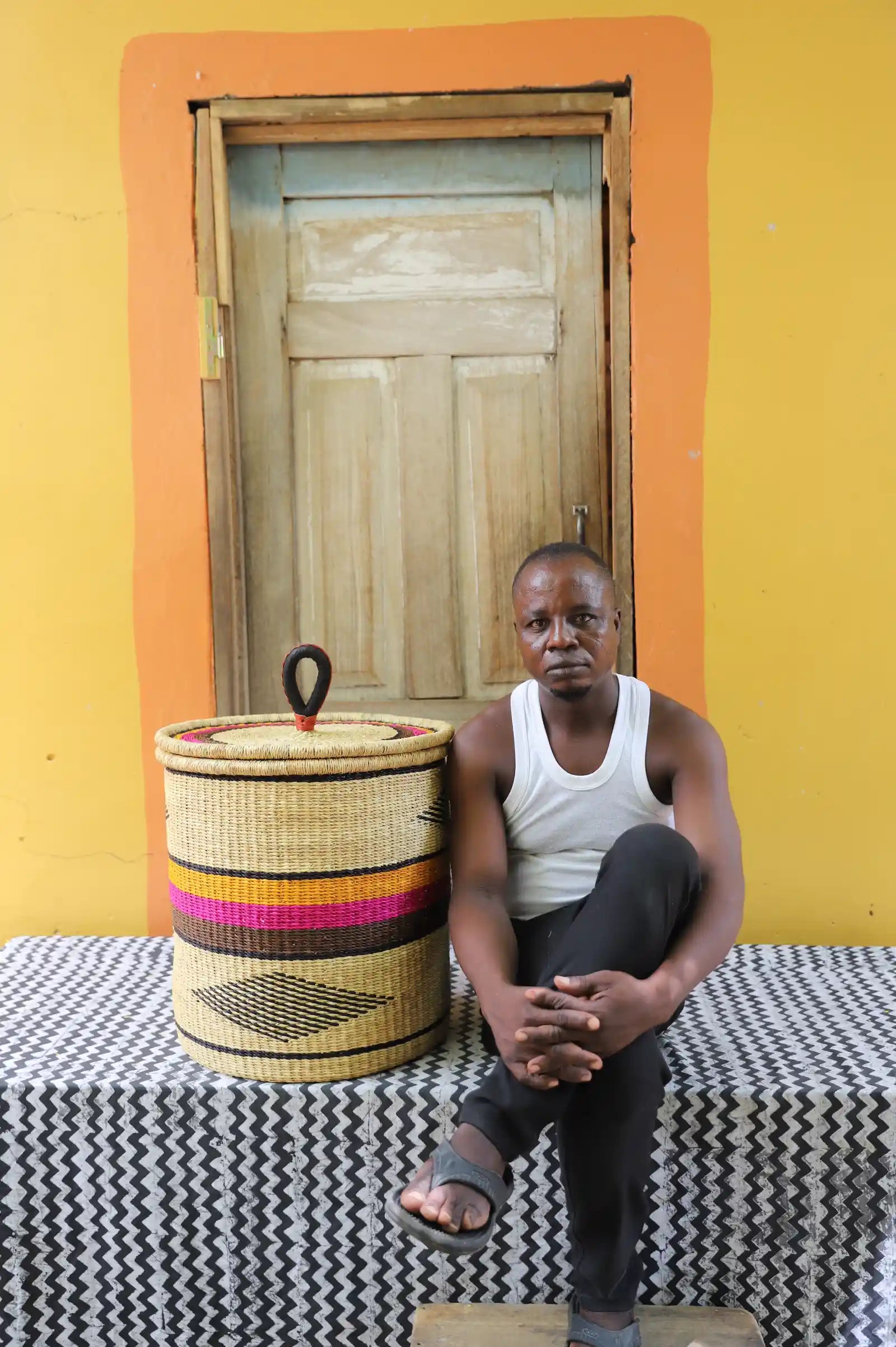 Handwoven Bolga laundry basket with pink and orange bands, beside a seated man in a white vest.