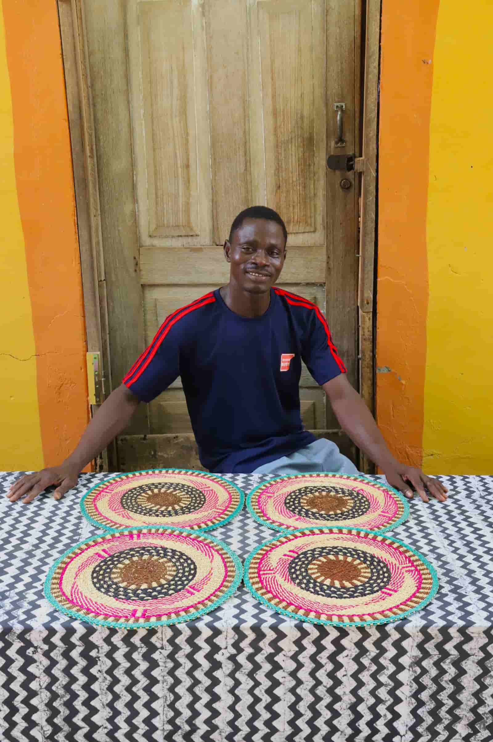 Artisan sitting with woven placemats on a black and white cloth against a wooden door.