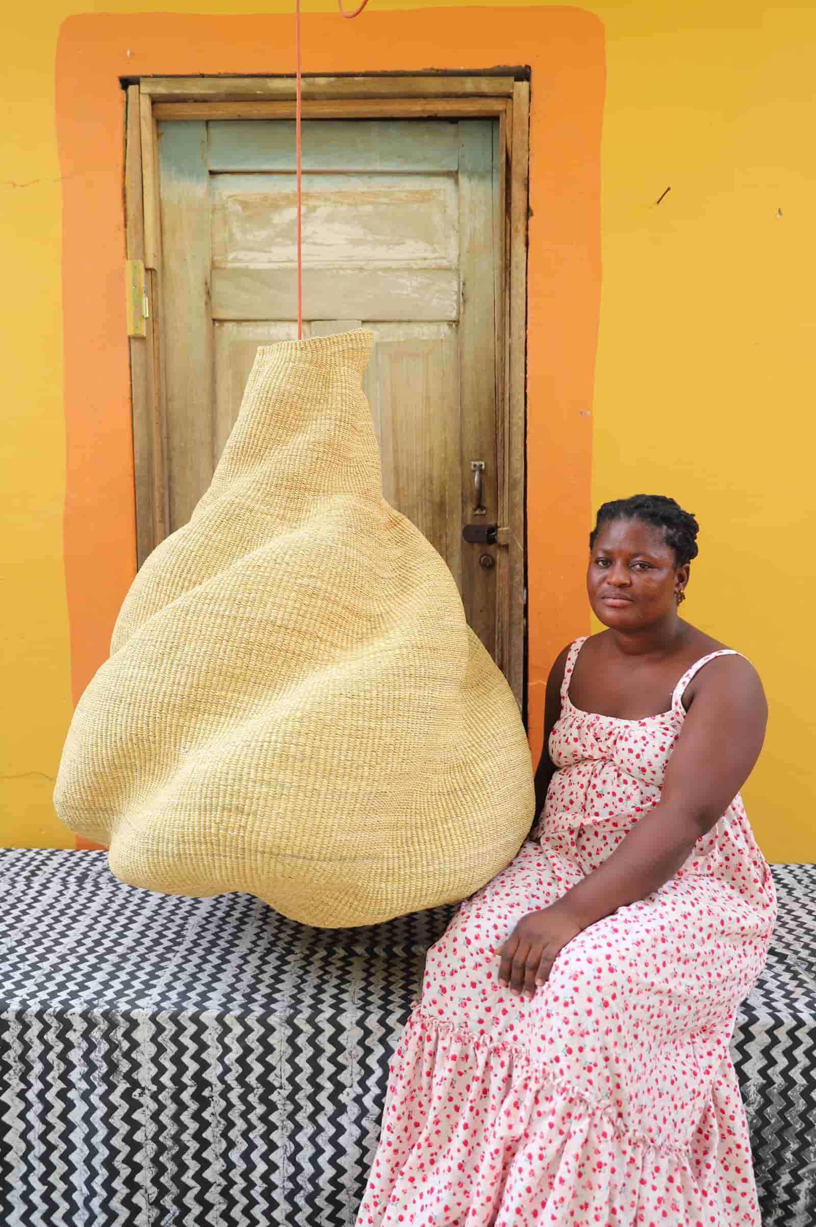 Woman in a floral dress sitting next to a large woven basket against a yellow wall.