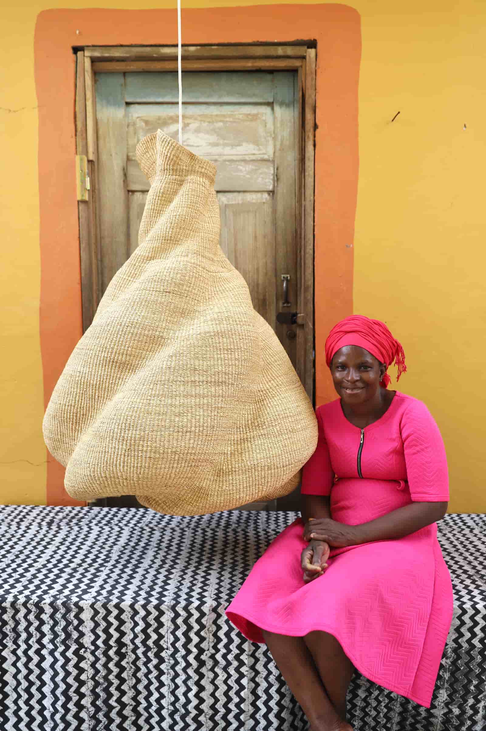 Woman in a pink dress sitting next to a large woven basket against a yellow wall.