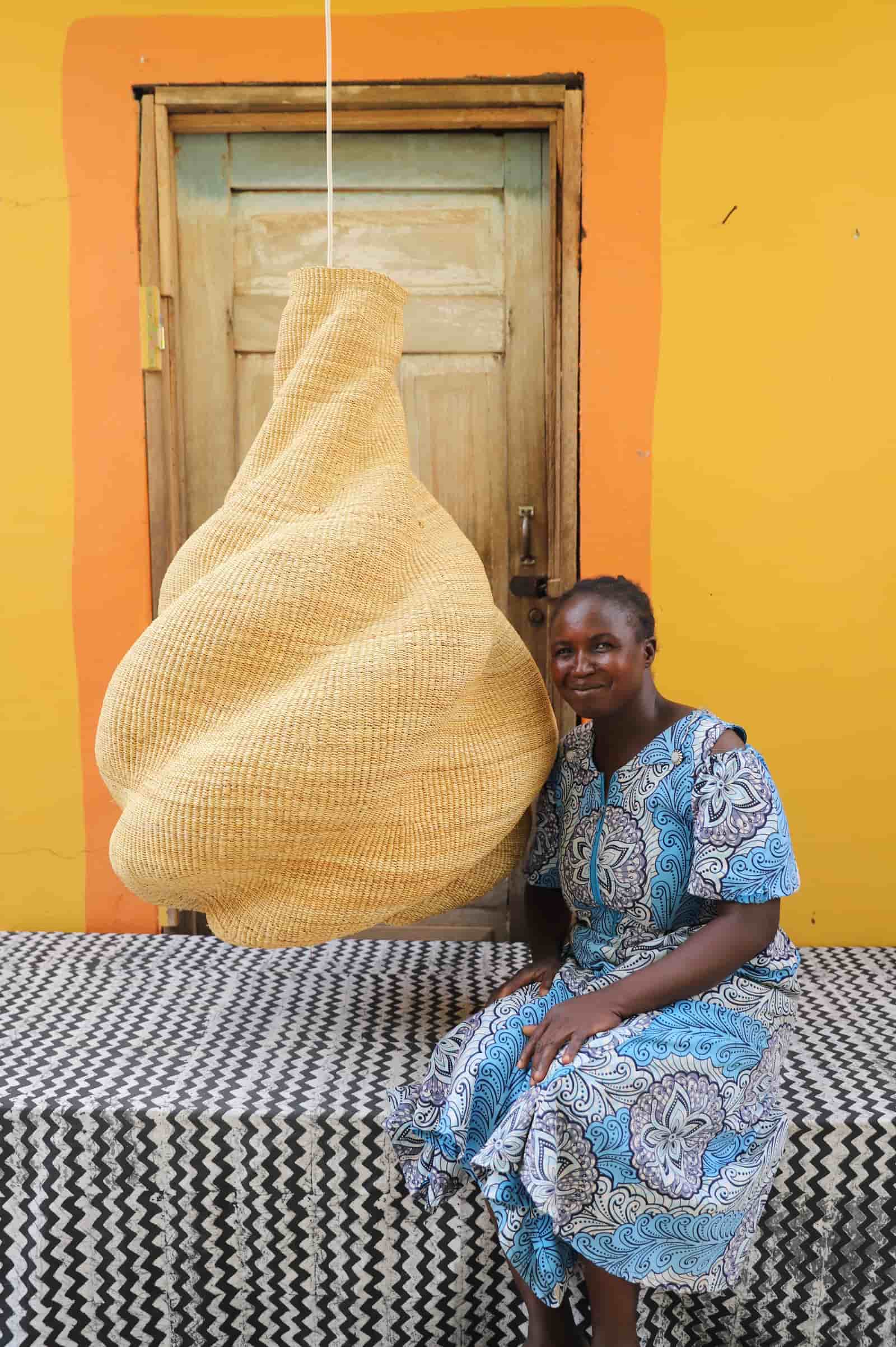 Woman in a blue patterned dress sitting next to a large woven object against a yellow wall.