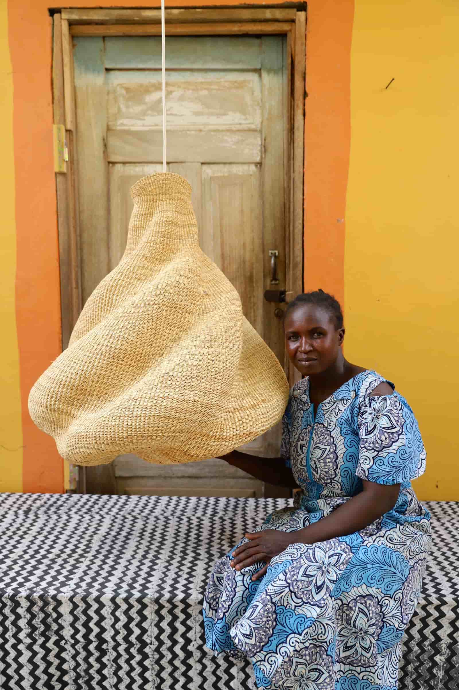 Woman sitting on a bed with a woven basket over her head against a yellow wall.