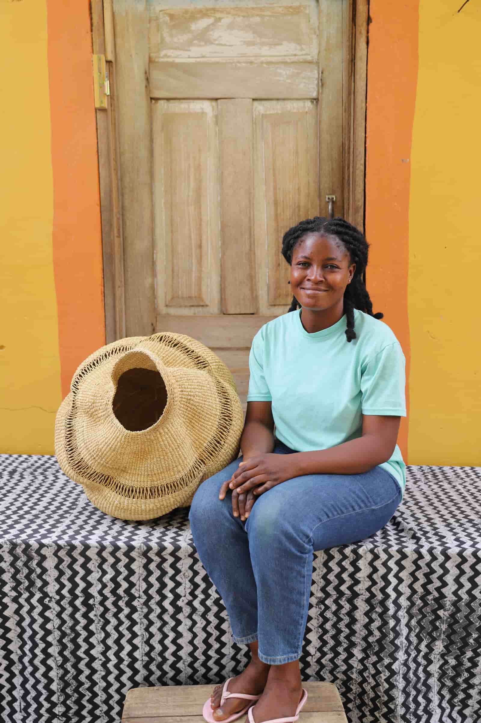 Woman sitting next to a woven conch-shaped lampshade on a black and white patterned cloth with a wooden door in the background.