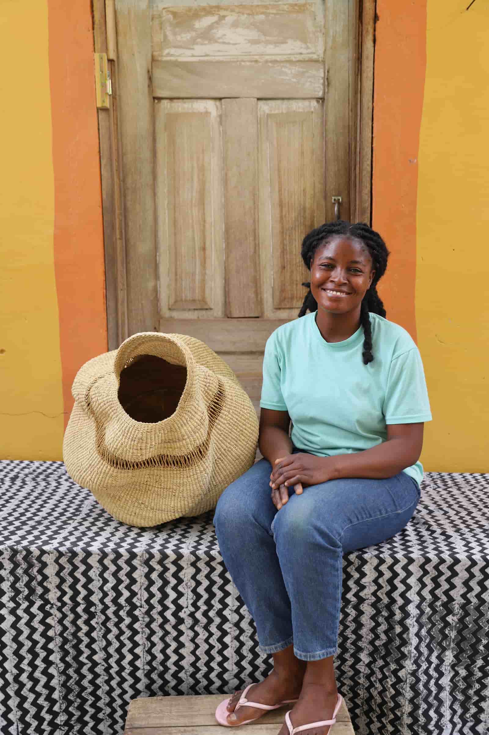Woman sitting next to a woven conch-shaped lampshade on a black and white patterned cloth with a wooden door in the background.