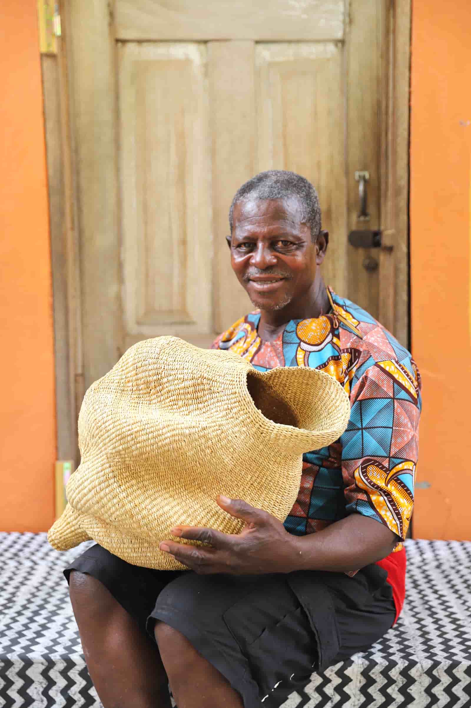 Man  holding a woven conch lampshade on a black and white patterned tablecloth with a wooden door in the background.