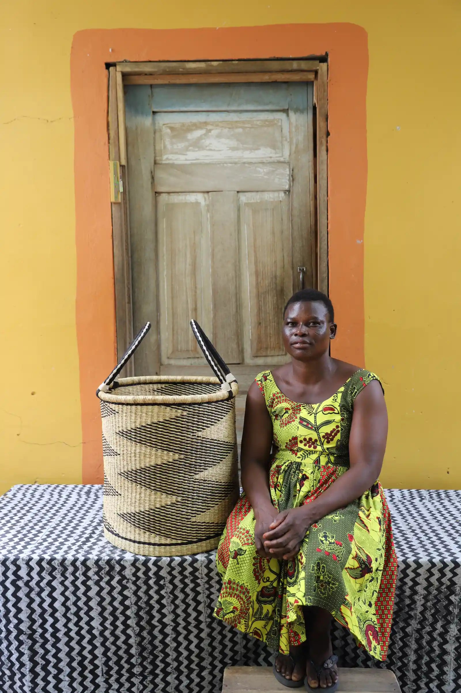 Medium handwoven Bolga laundry basket in natural with black sawtooth stripes, beside a seated woman in green.