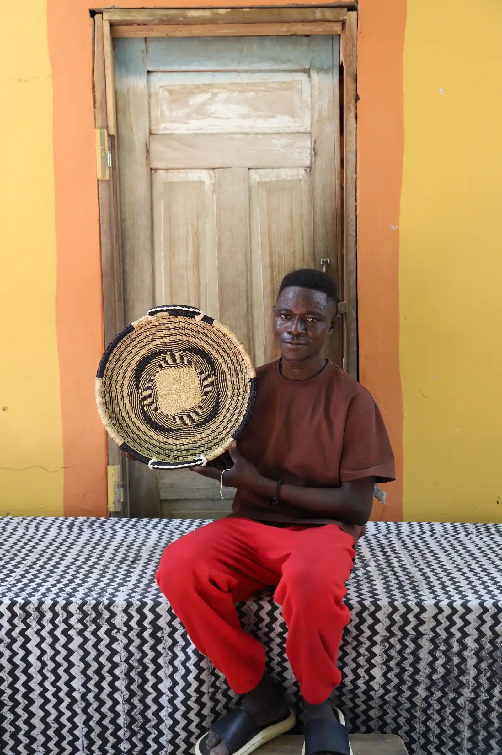 Handwoven Bolga wall tray disc in natural with yellow centre, held by a seated man in red.