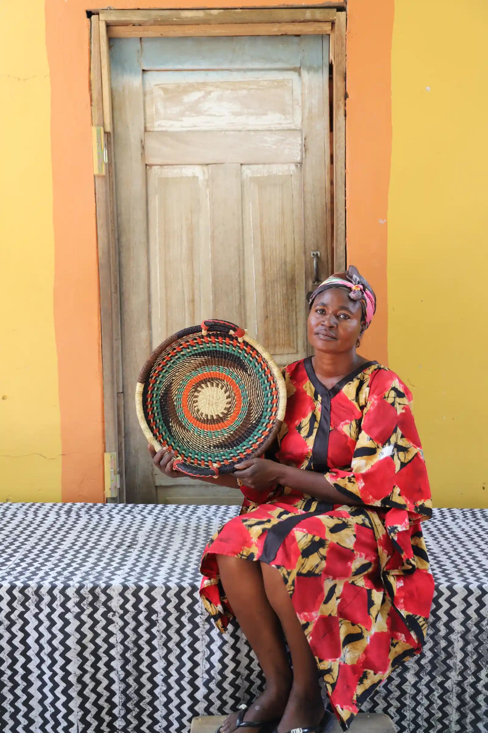 Handwoven Bolga wall tray disc with green and orange bands, held by a seated woman in red floral dress.