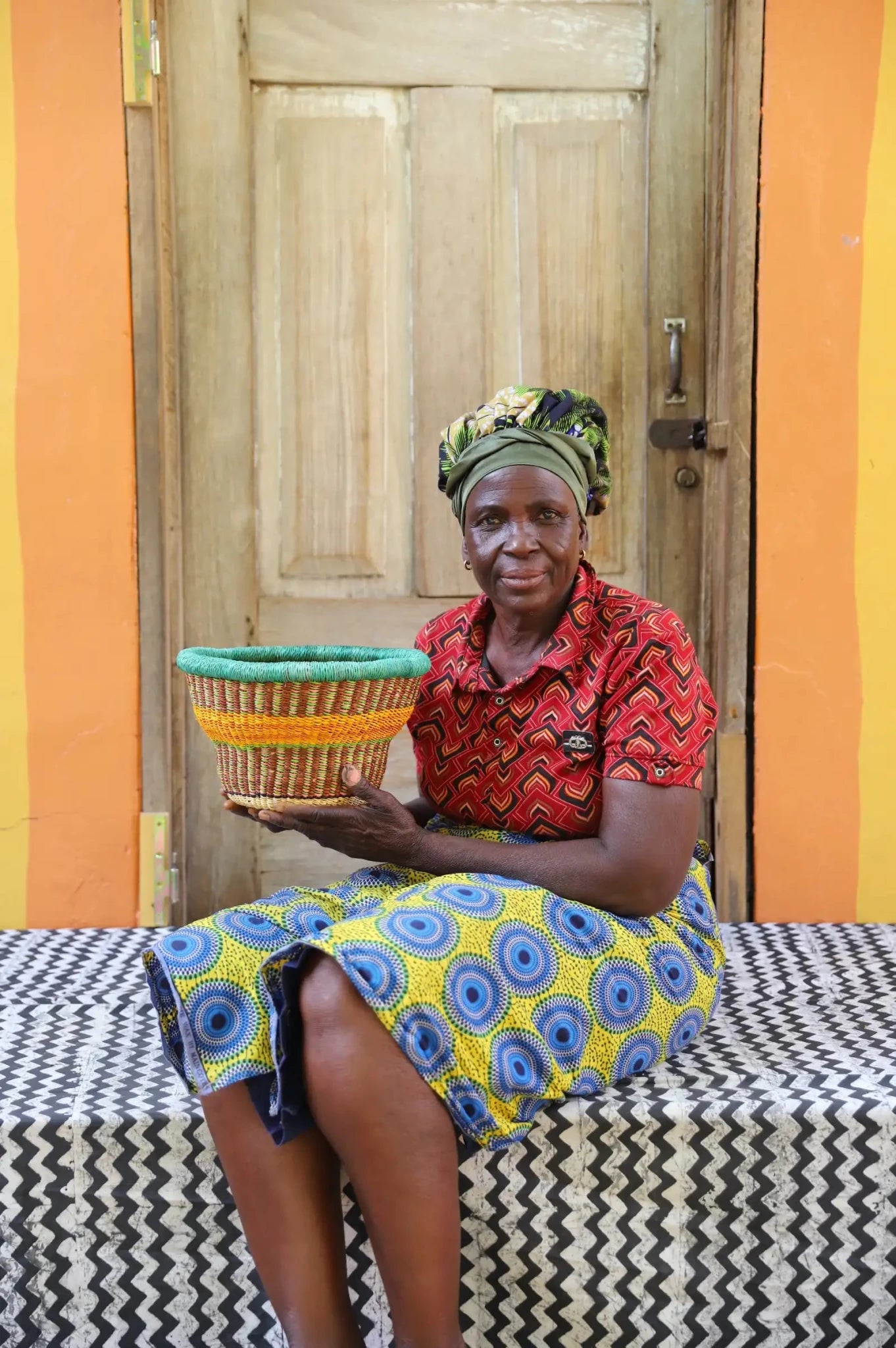 Woman holding a woven basket in front of a wooden door with colorful walls.