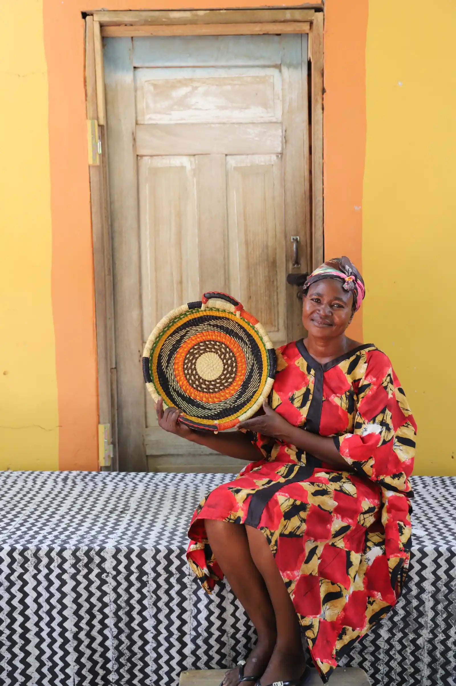 Handwoven Bolga wall tray disc with orange and black bands, held by a seated woman in red floral.