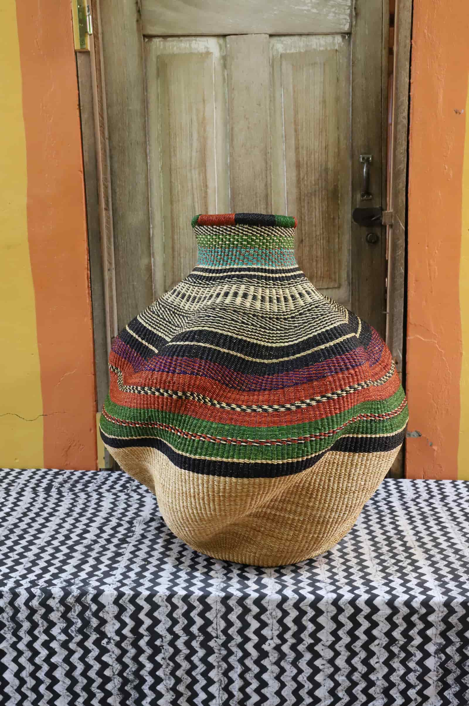 Large handwoven De-engo art basket with a natural straw base and terracotta, green and black stripes, on a chevron cloth.