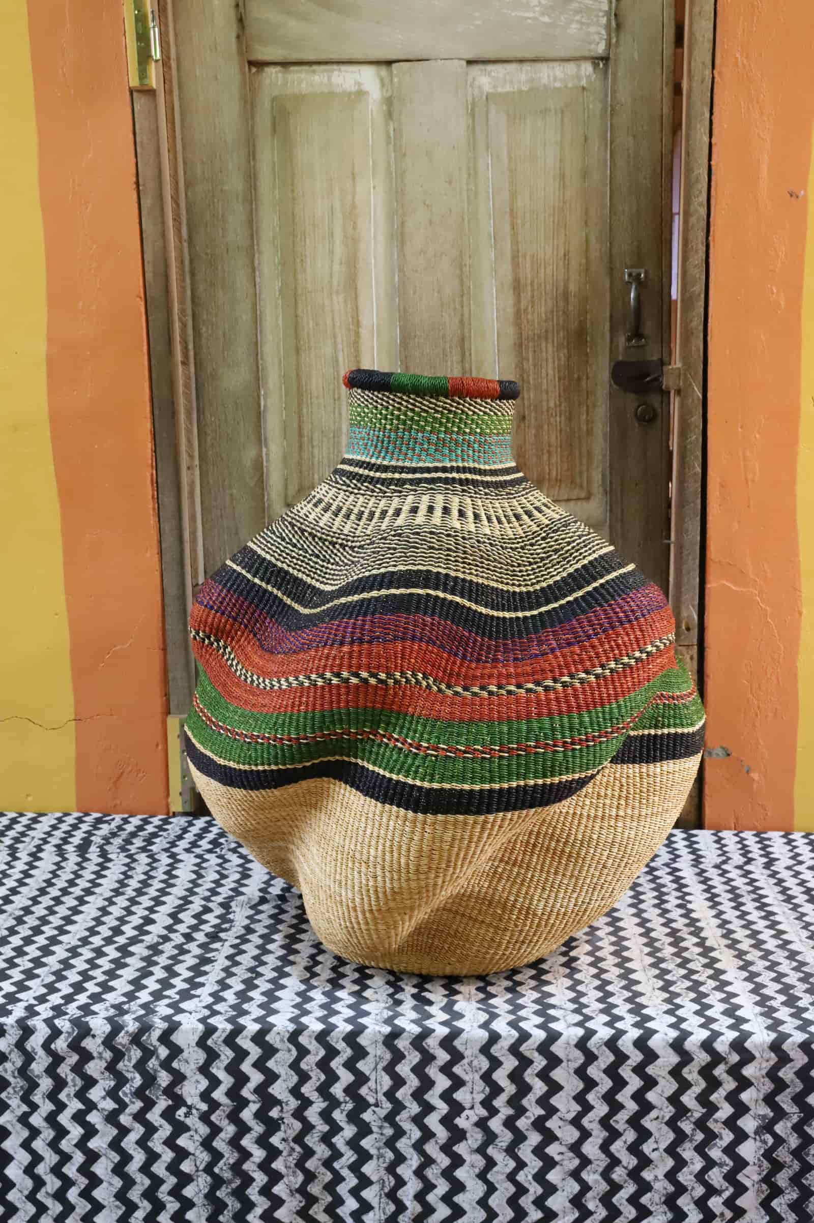 Large handwoven De-engo art basket in natural straw with terracotta, green and black band stripes, on a chevron cloth.