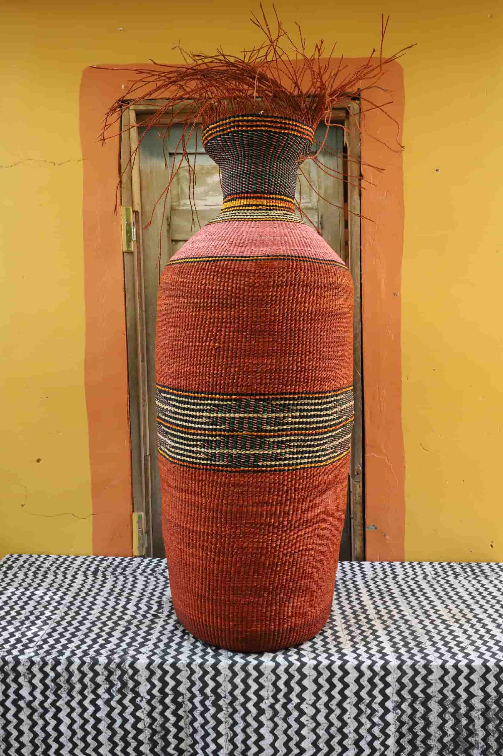 Tall Bolga art basket in terracotta with black and straw banded stripes, holding dried branches, on a chevron cloth.