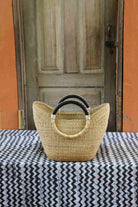 Woven handbag with black handles on a tablecloth in front of a wooden door.