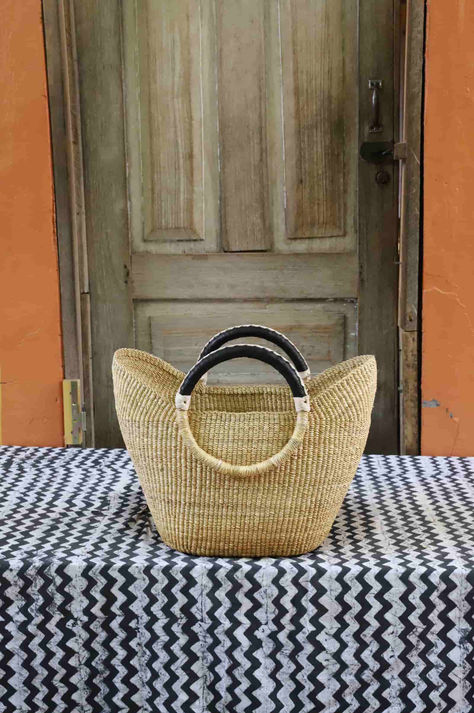 Woven handbag with black handles on a tablecloth in front of a wooden door.