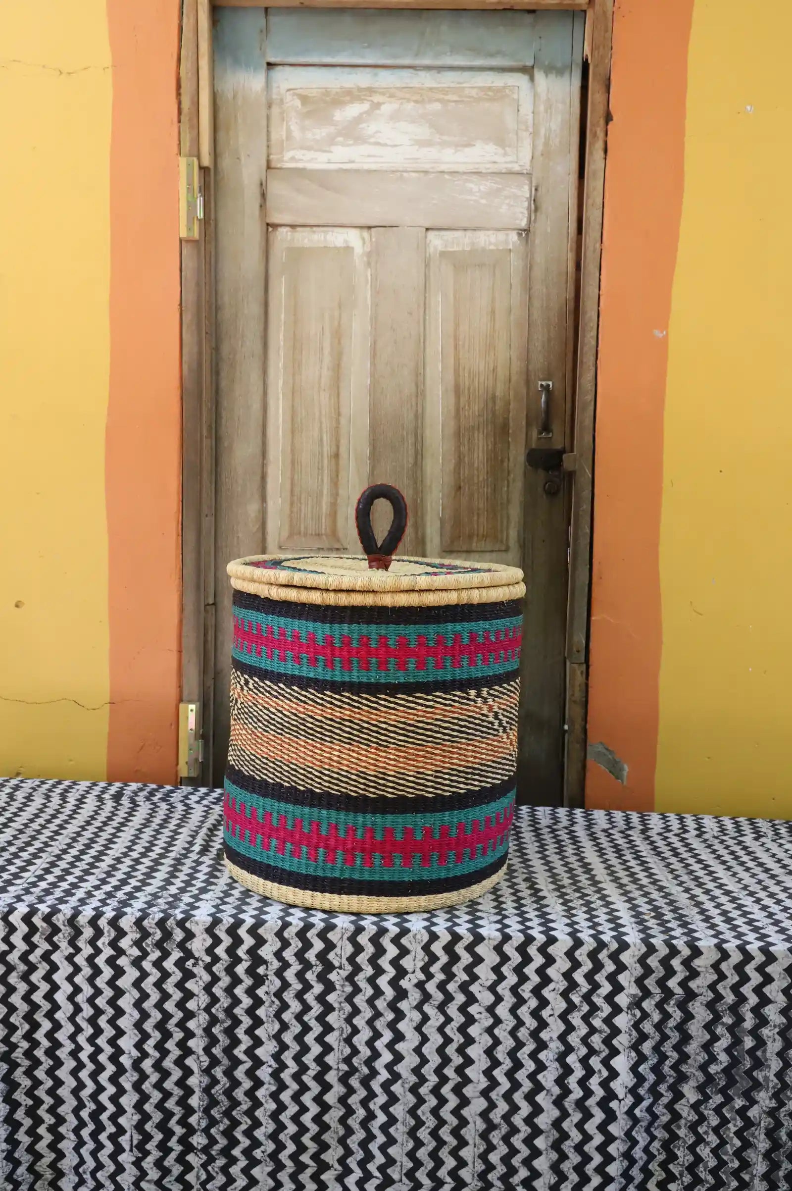 Handwoven Bolga laundry basket with coiled rim and lid in natural straw with fine pink, black and orange bands.