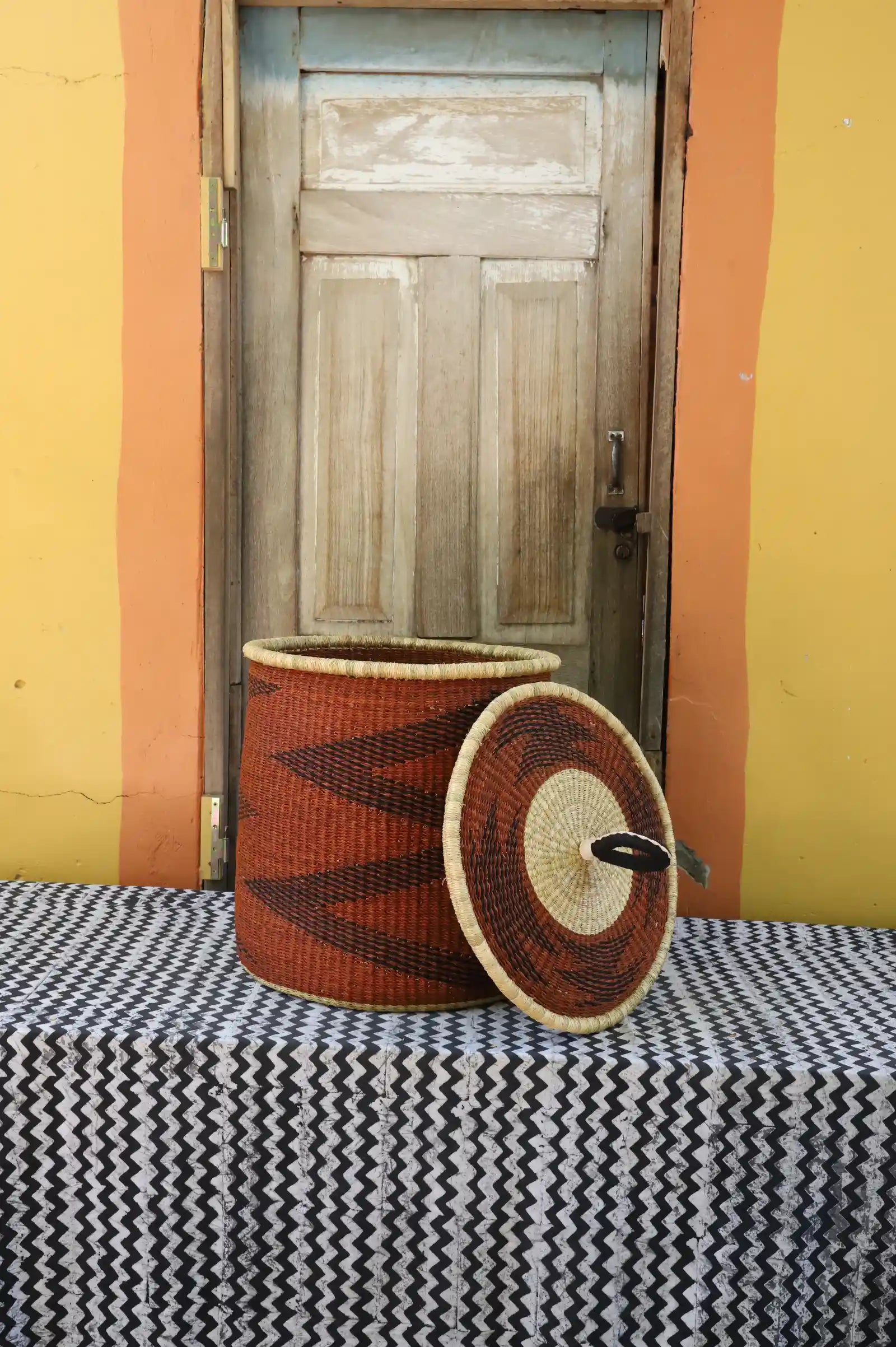 Handwoven Bolga laundry basket with lid removed, rust-orange body with dark zigzag and natural rim lid.