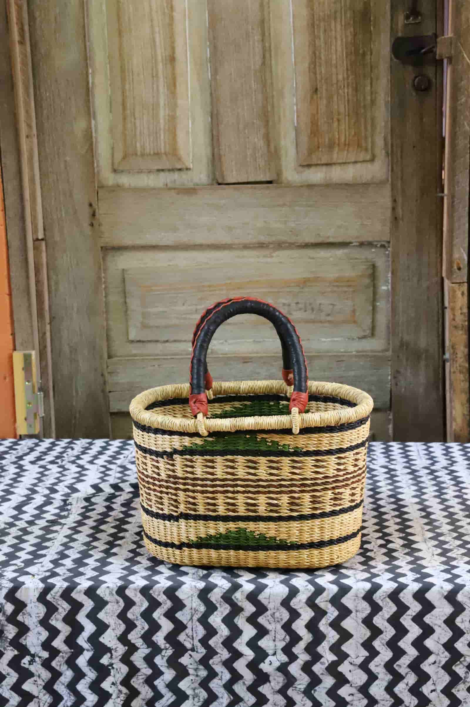 Small oval woven carry basket in natural straw with black and green stripes, leather-wrapped handle, against a wooden door