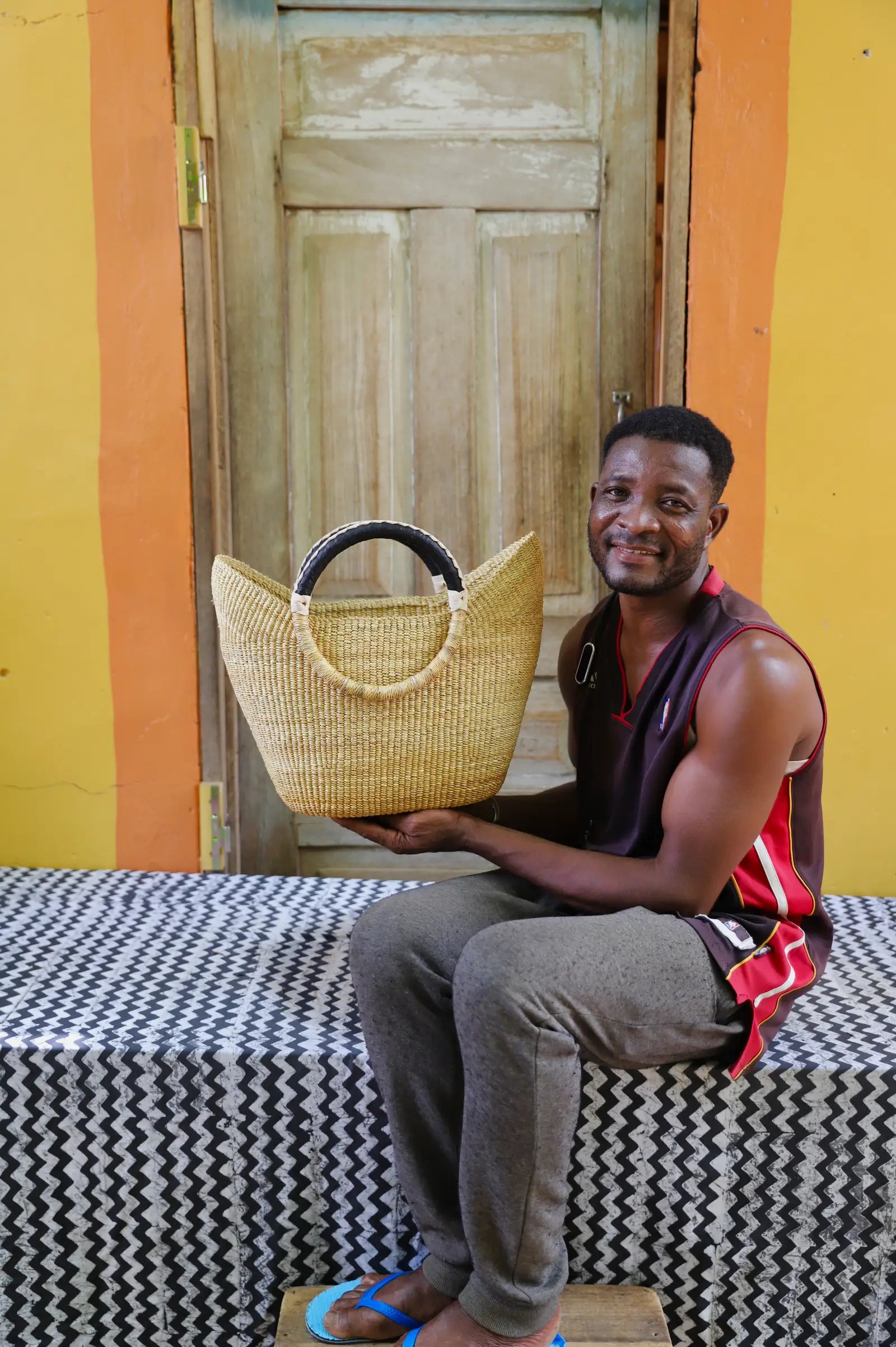 Man holding a woven basket in front of a yellow wall and wooden door.