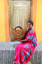 Woman in a bright pink dress with blue patterns holding a woven basket against a colorful wall.