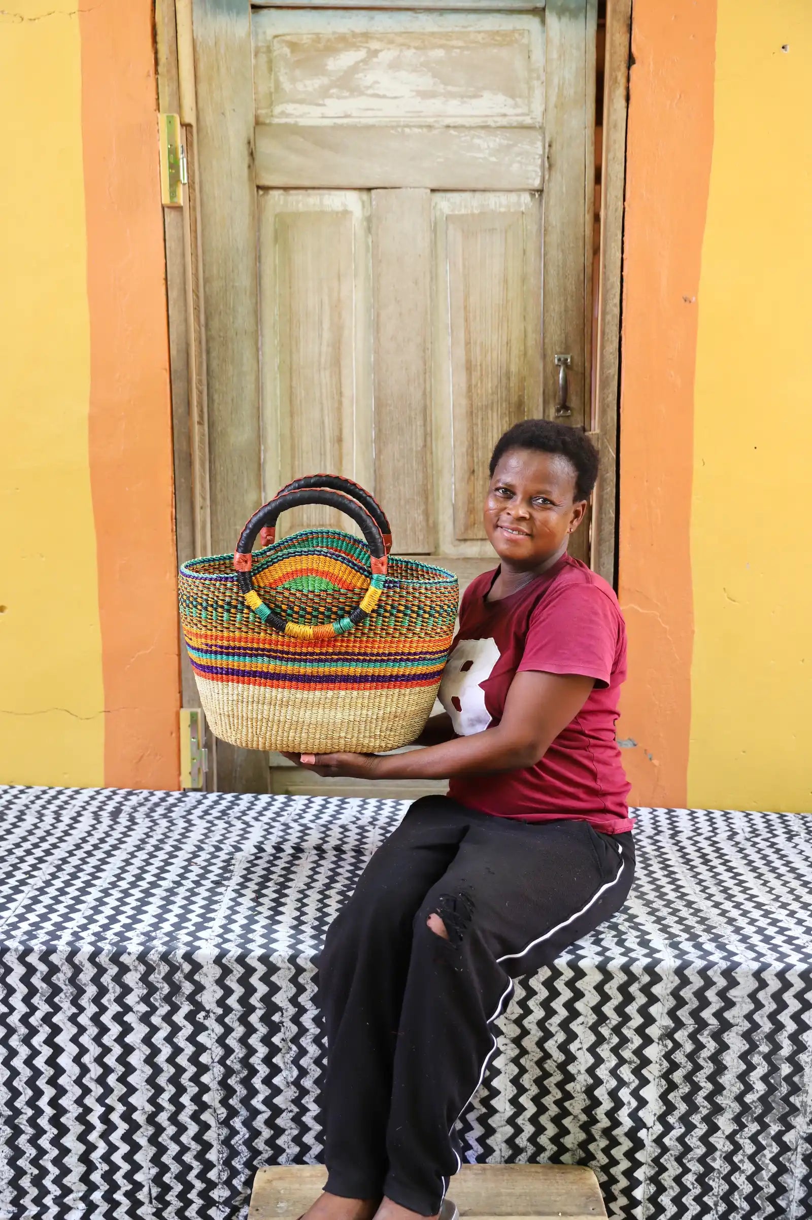 Person holding a colorful woven basket in front of a wooden door with a yellow wall.
