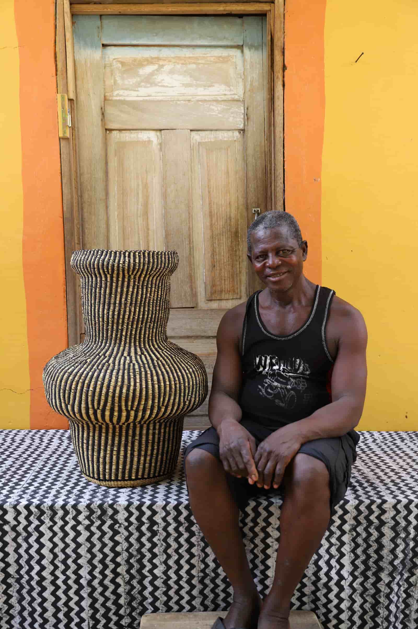 Man sitting next to a woven basket on a black and white patterned surface with a wooden door in the background.