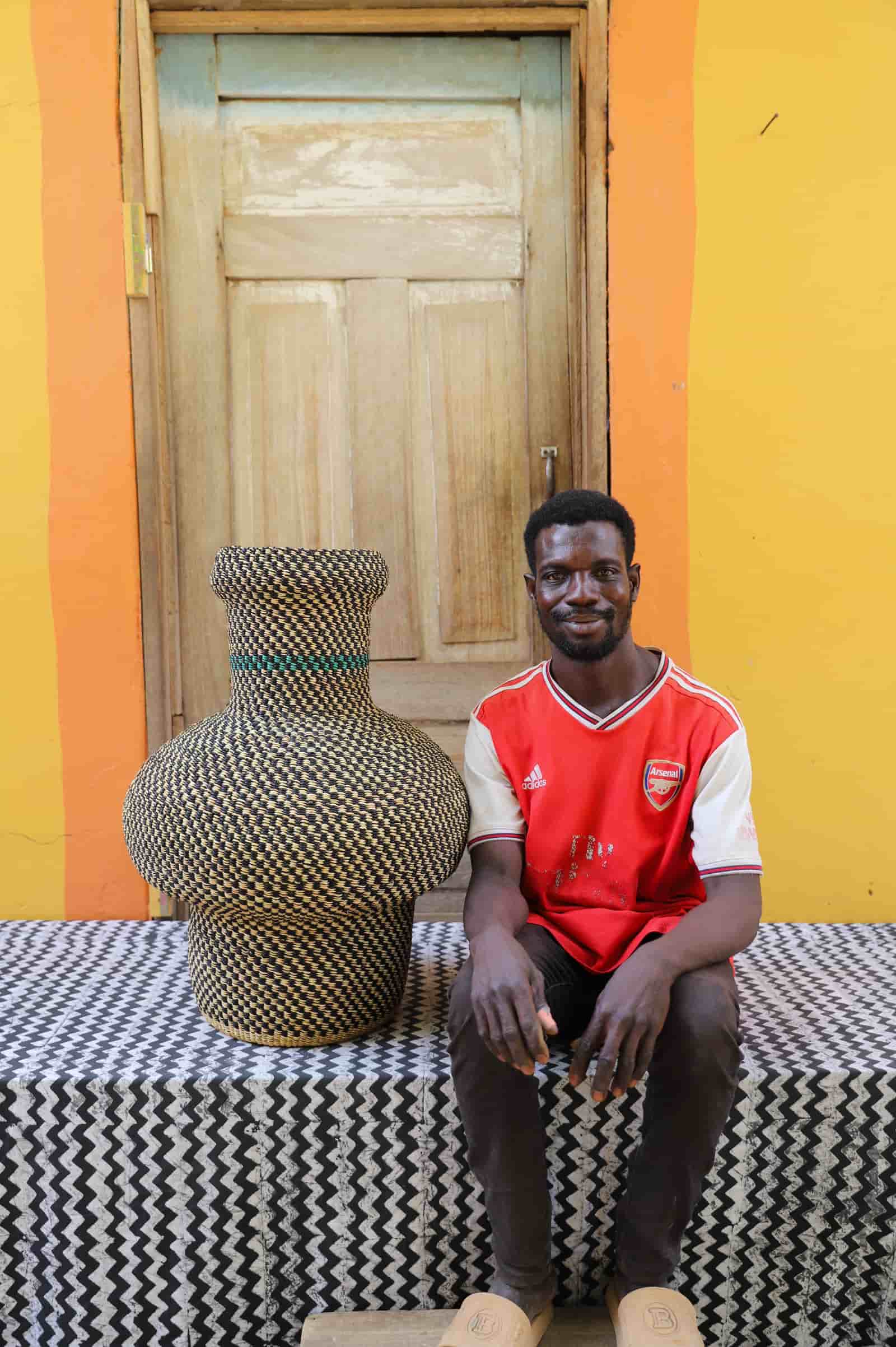 Man sitting next to a colorful woven basket on a black and white patterned surface with a wooden door in the background.