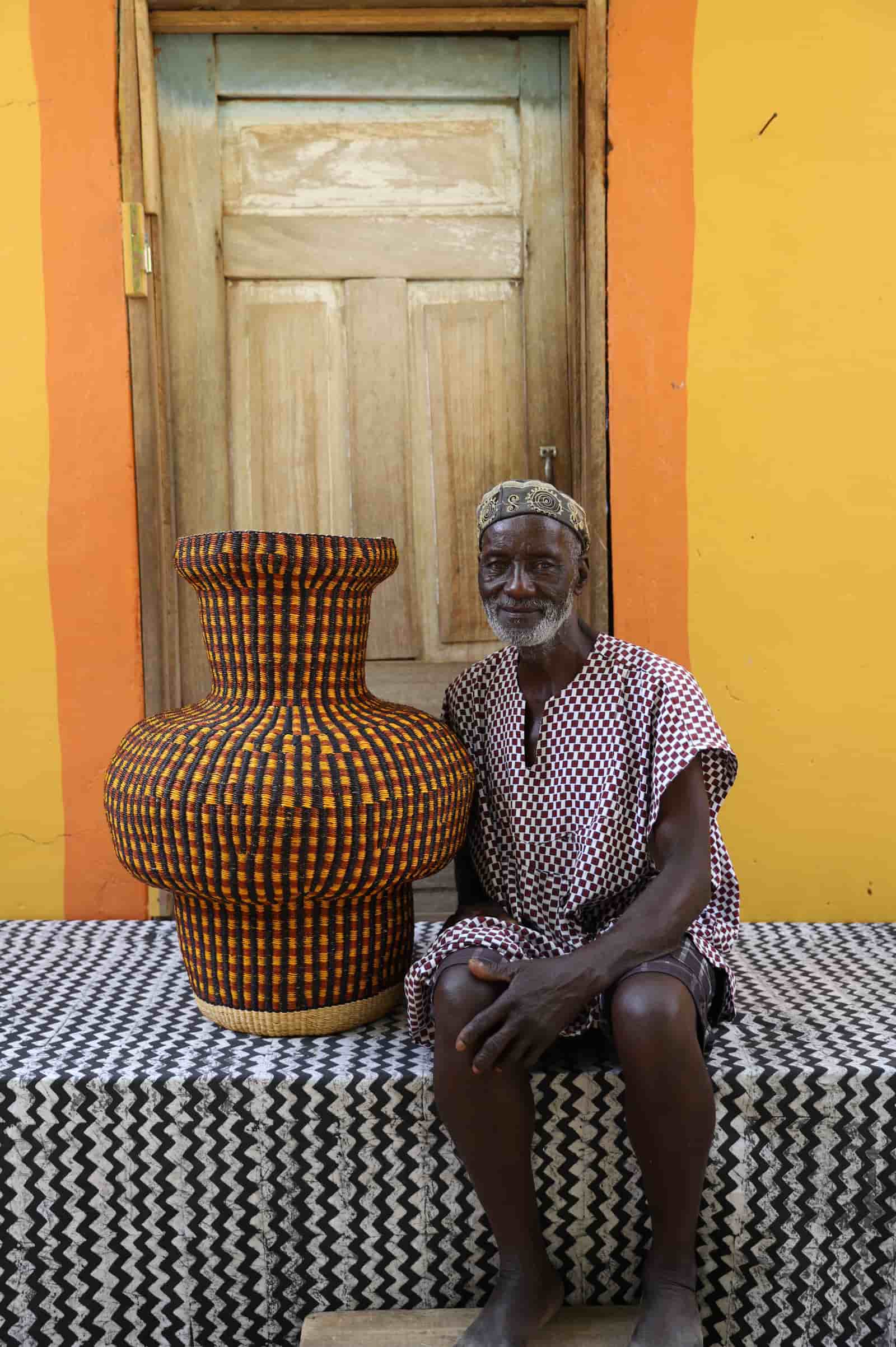 Man sitting next to a colorful woven basket on a black and white patterned surface with a wooden door in the background.