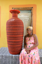 Woman in a pink dress sitting next to a large red woven vase against a yellow wall.