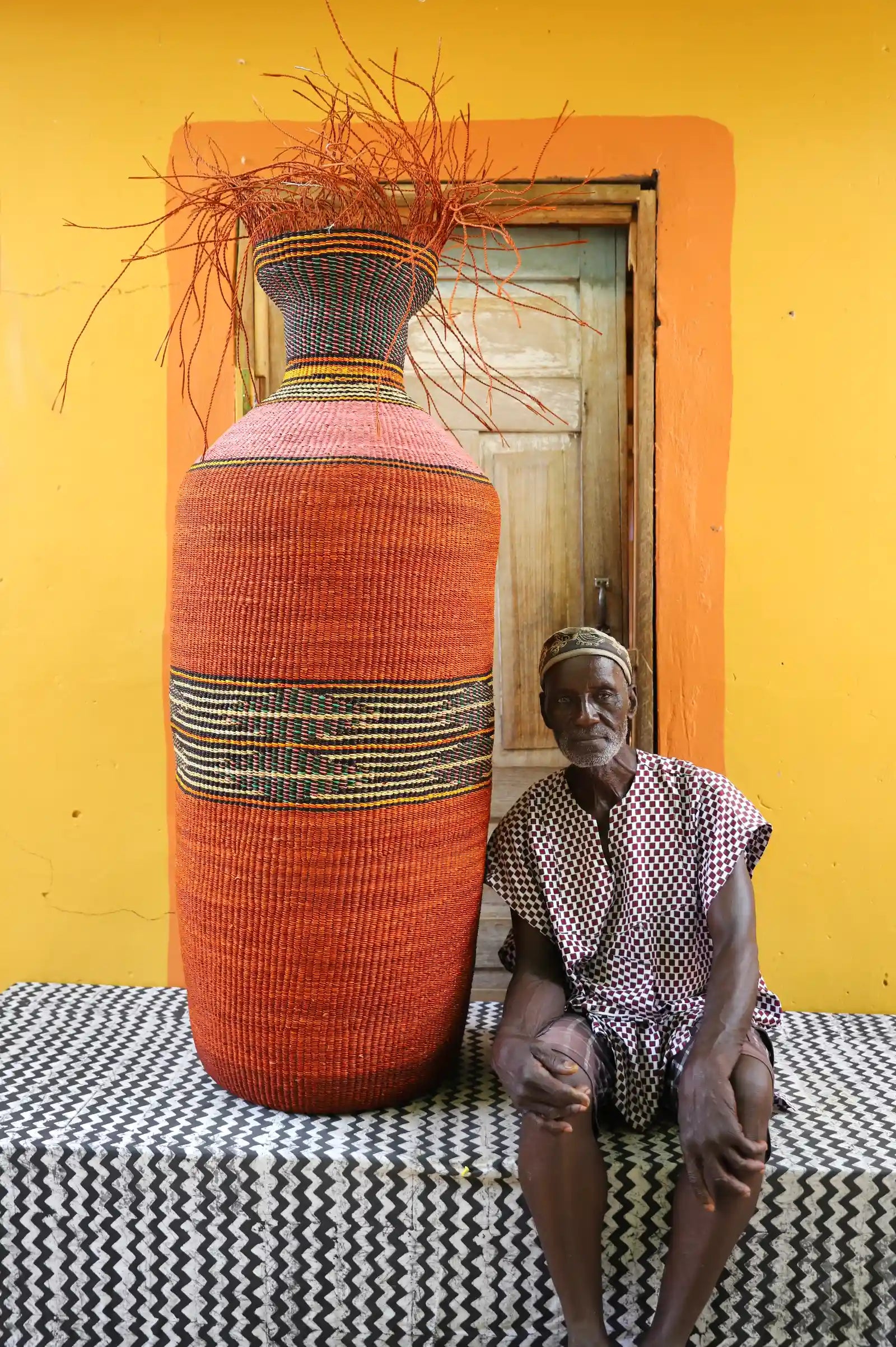 Man seated beside a tall terracotta Bolga art basket with black and straw banded stripes, against a yellow wall.