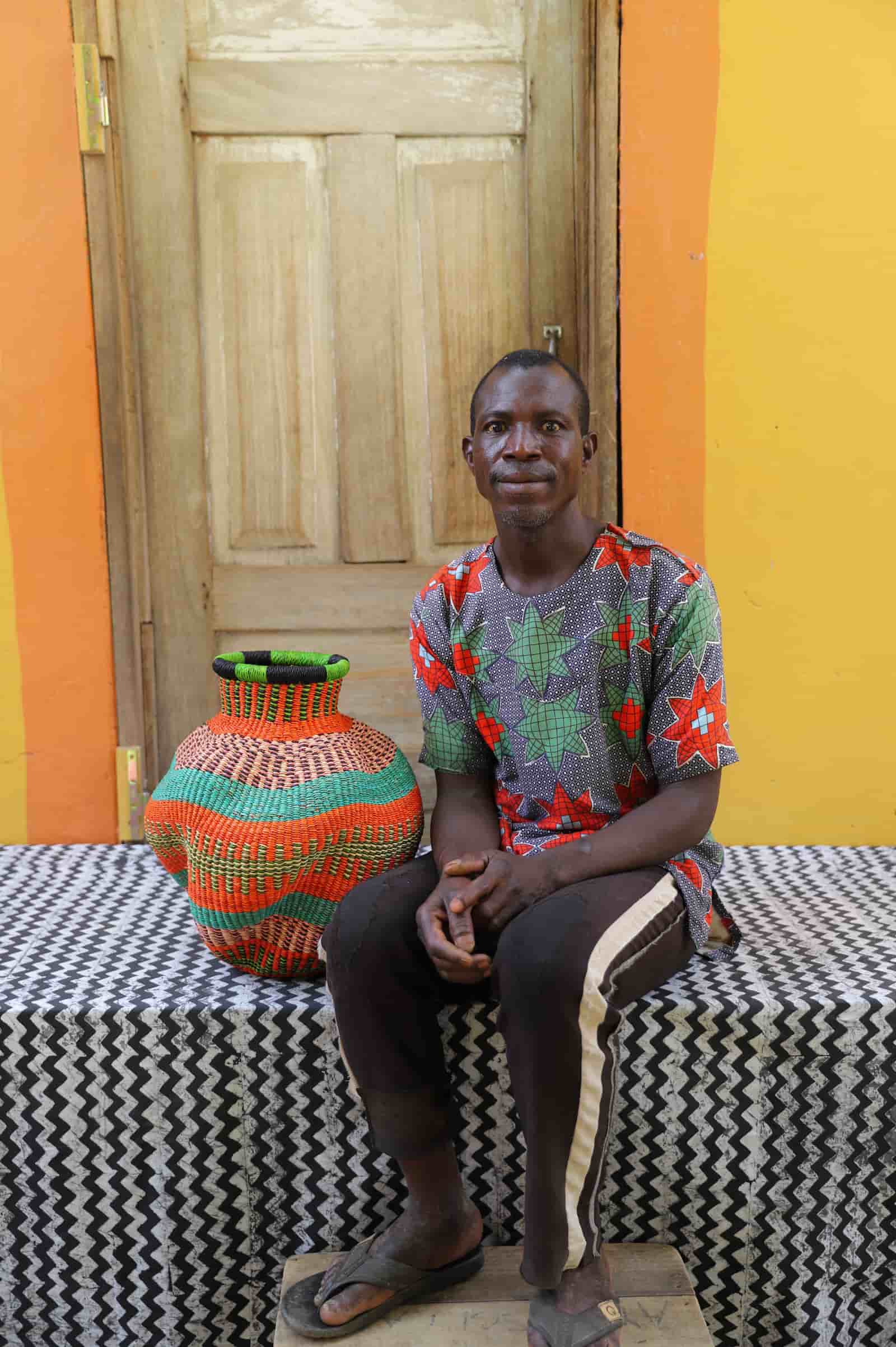 Man sitting on a patterned bench next to a large, colorful woven basket against a yellow wall.