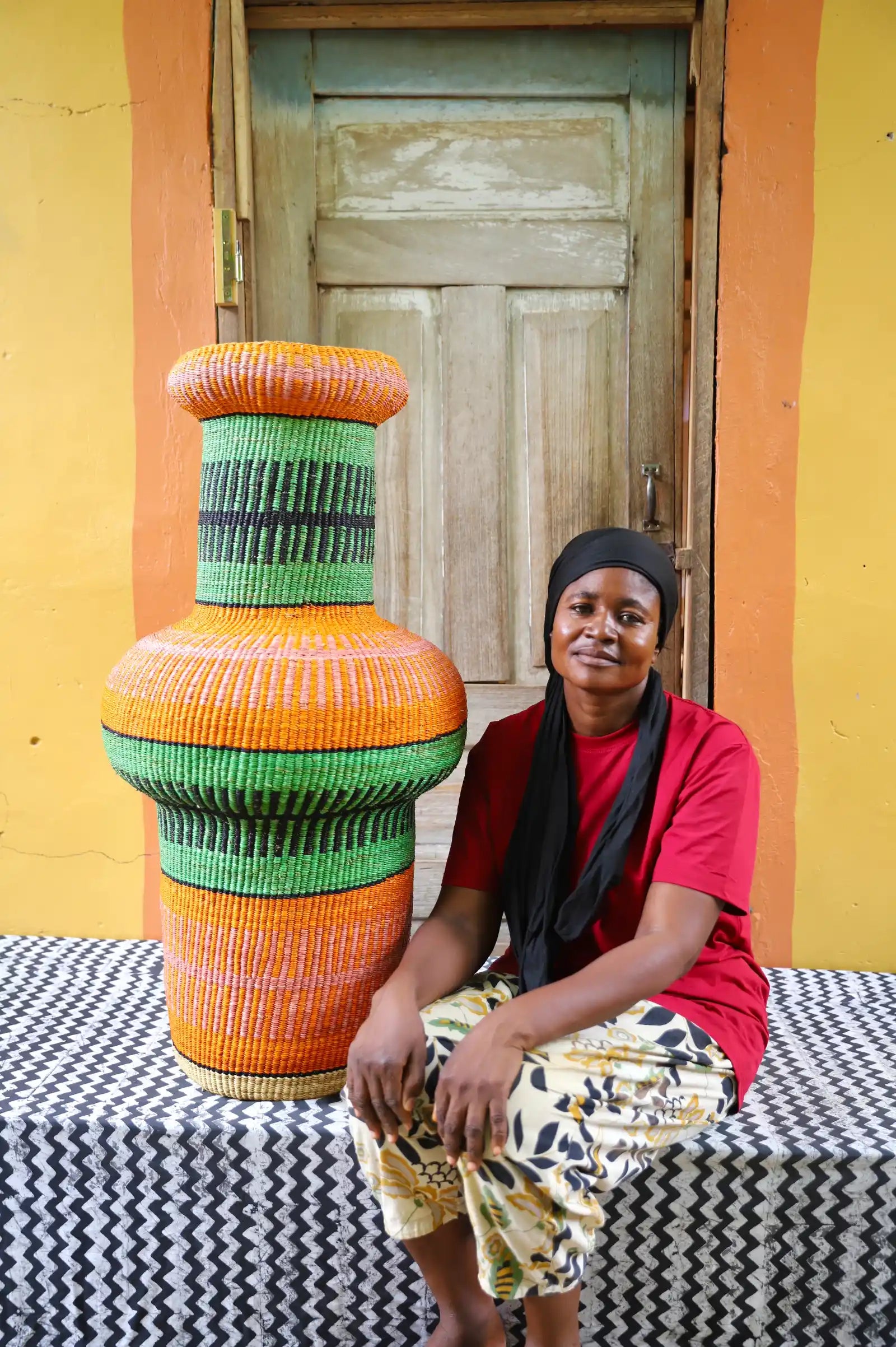 Woman sitting next to a colorful, woven décor piece on a black and white cloth against a wooden door.
