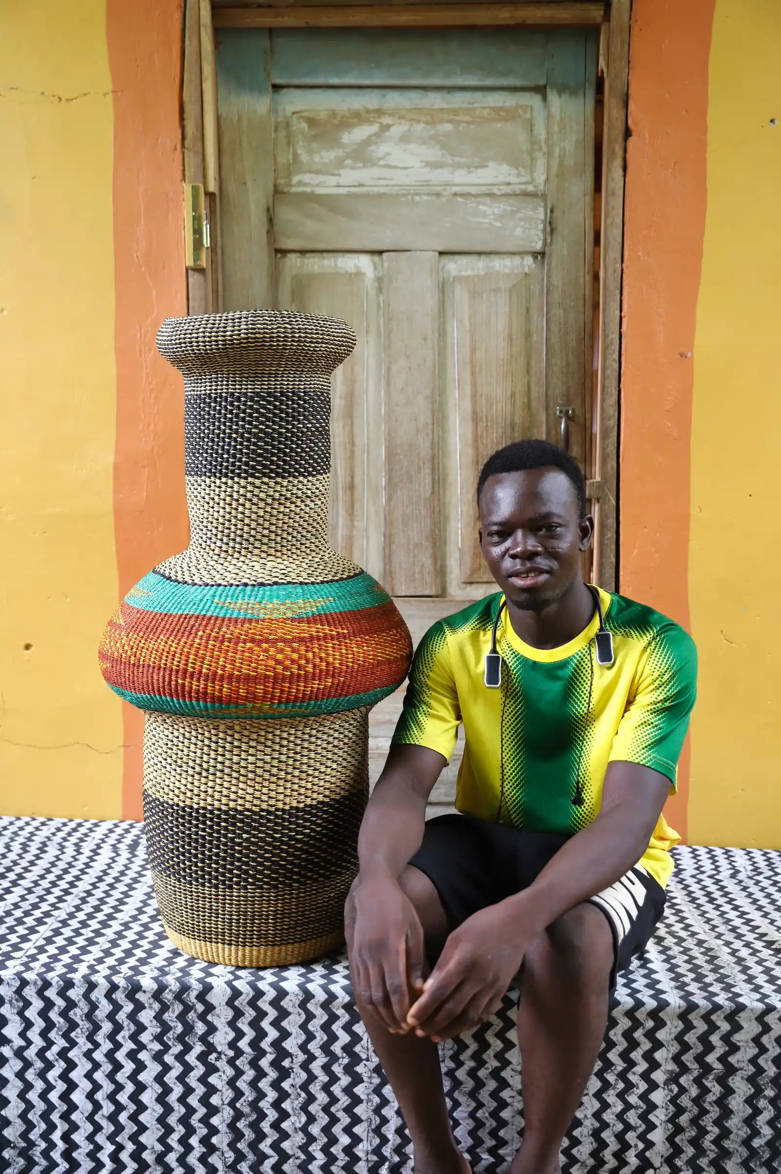 Man sitting next to a colorful, woven décor piece on a black and white cloth against a wooden door.