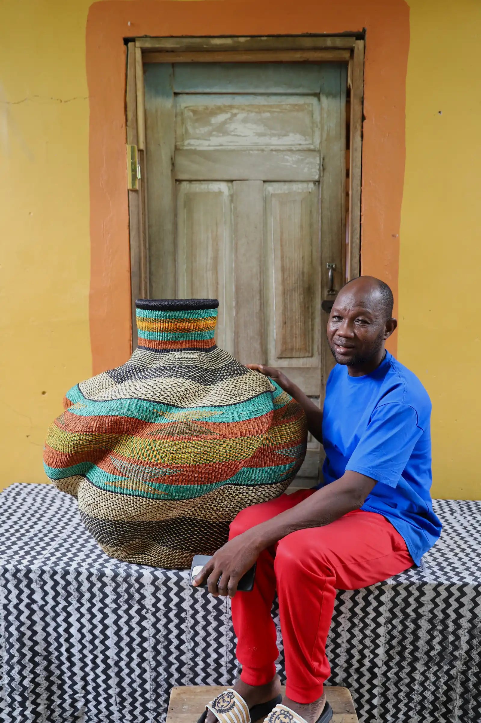 Man sitting on a patterned bench next to a large, colorful woven basket against a yellow wall.