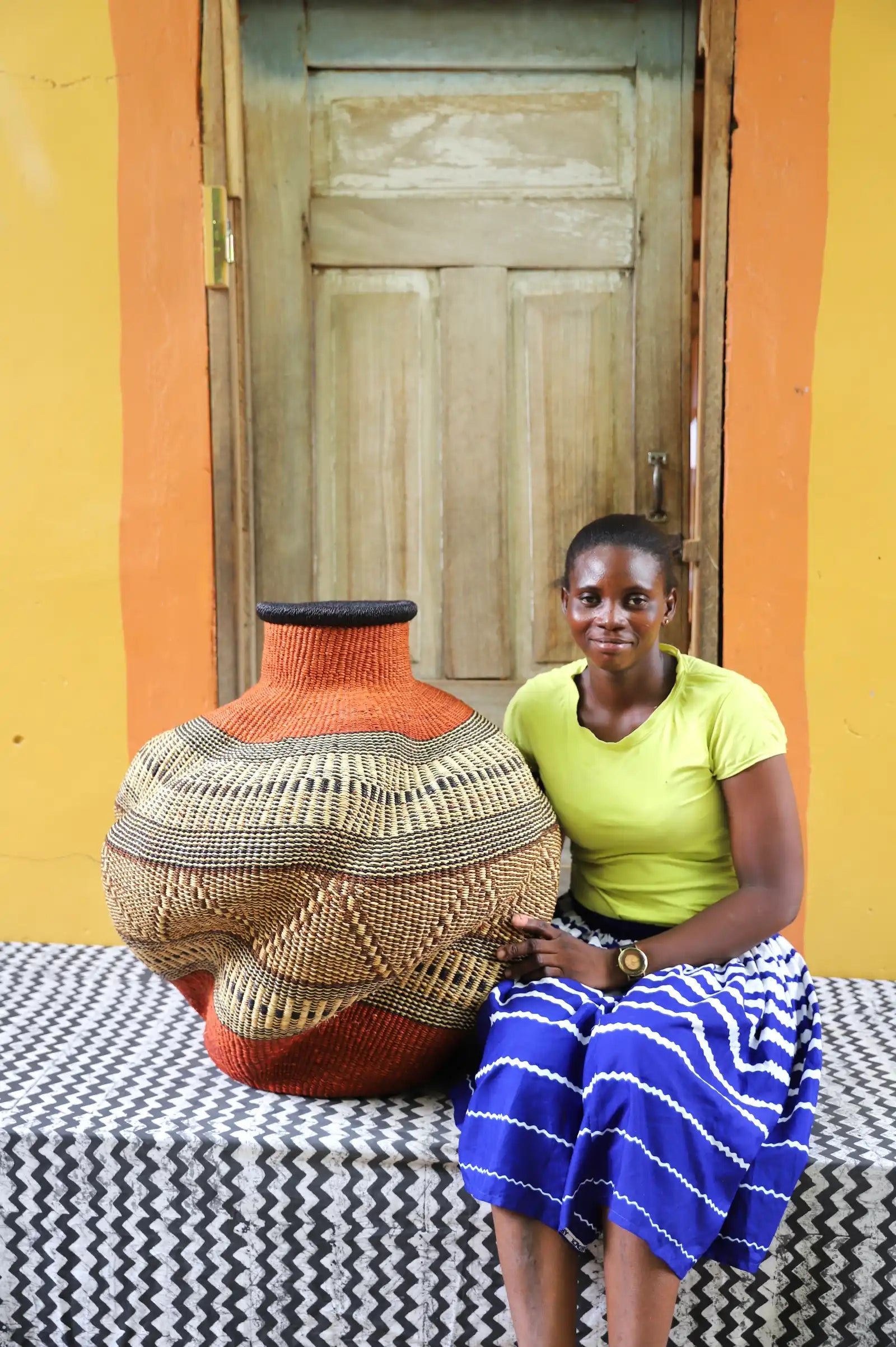Woman sitting on a patterned bench next to a large, colorful woven basket against a yellow wall.