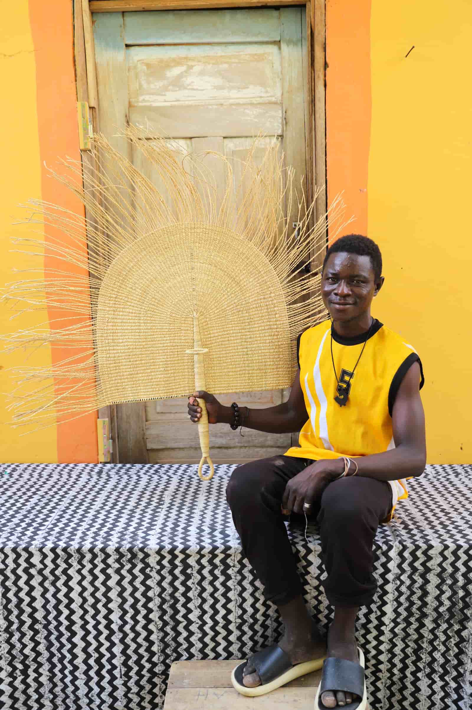 Man in a yellow sleeveless shirt sitting on a patterned bench against a bright yellow wall with a woven art piece.