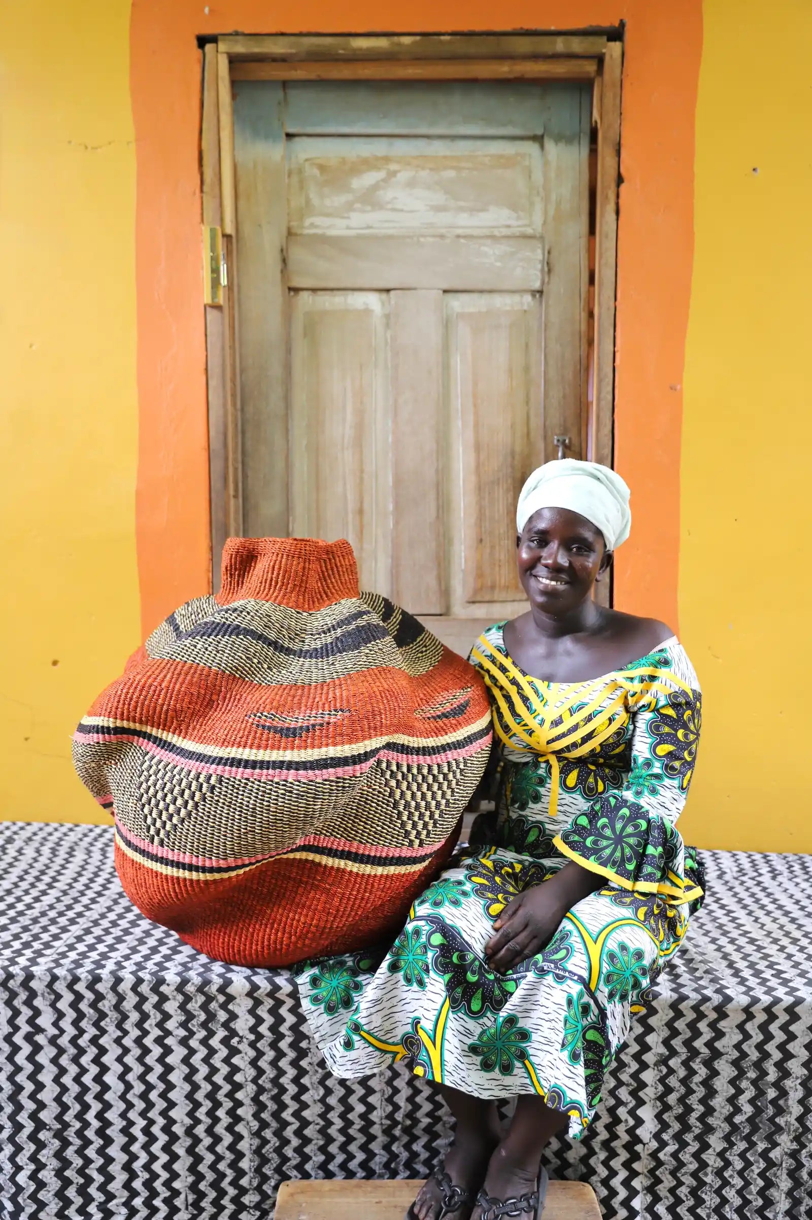 An artisan seated next to a handcrafted decorative basket  on a black and white cloth against a wooden door.