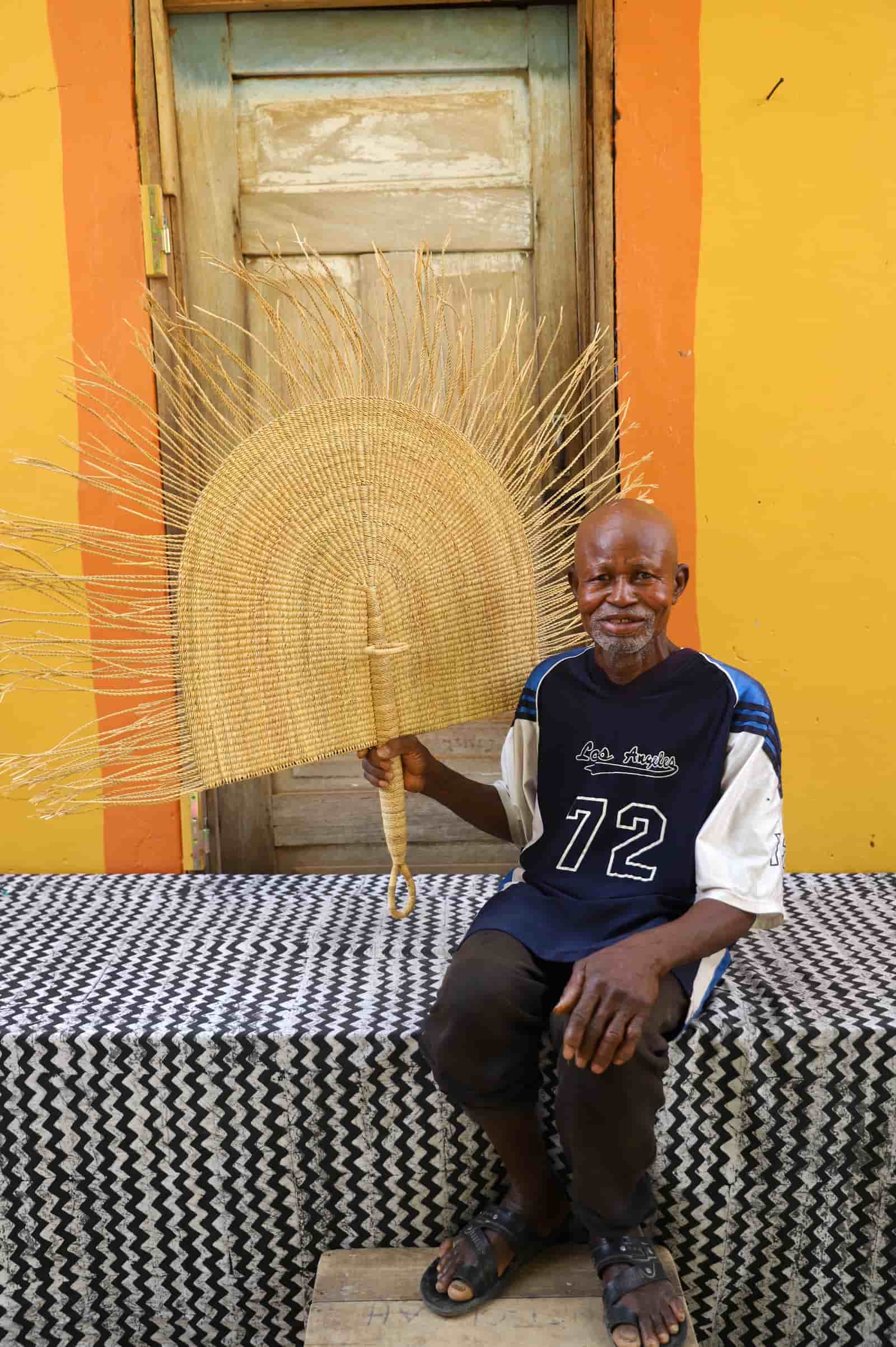 Man holding a large woven fan against a colorful wall.