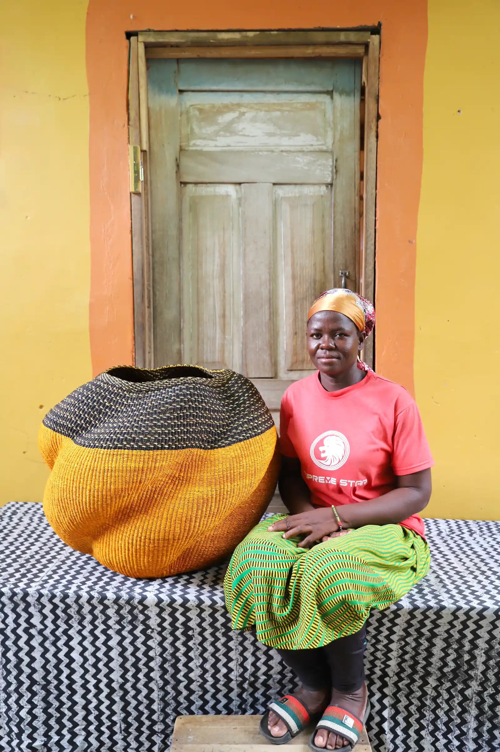 Woman seated beside an amber Bolga basket with a dark striped collar, against a yellow wall and wooden door.