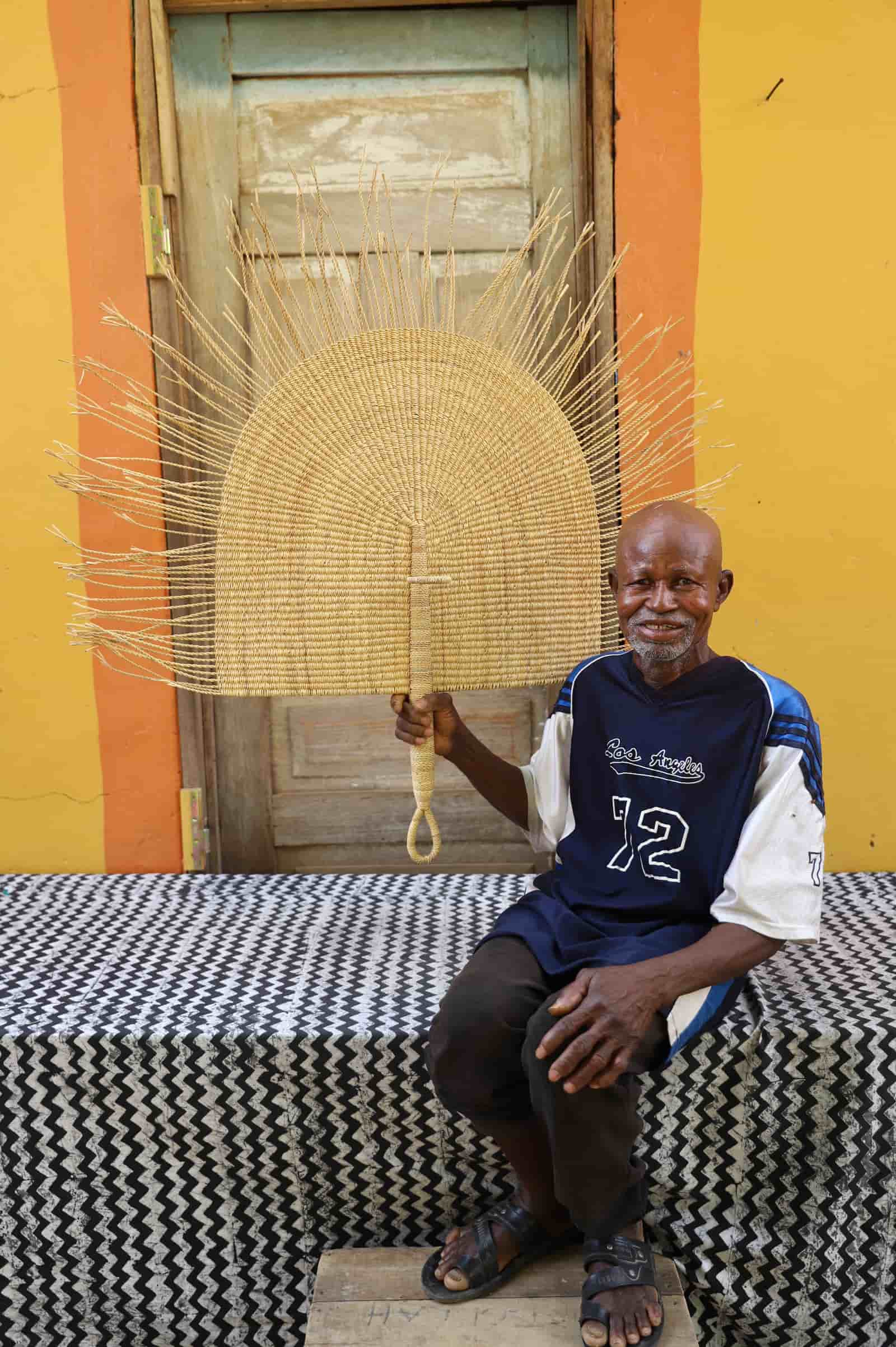 Man holding a woven fan against a colorful wall.