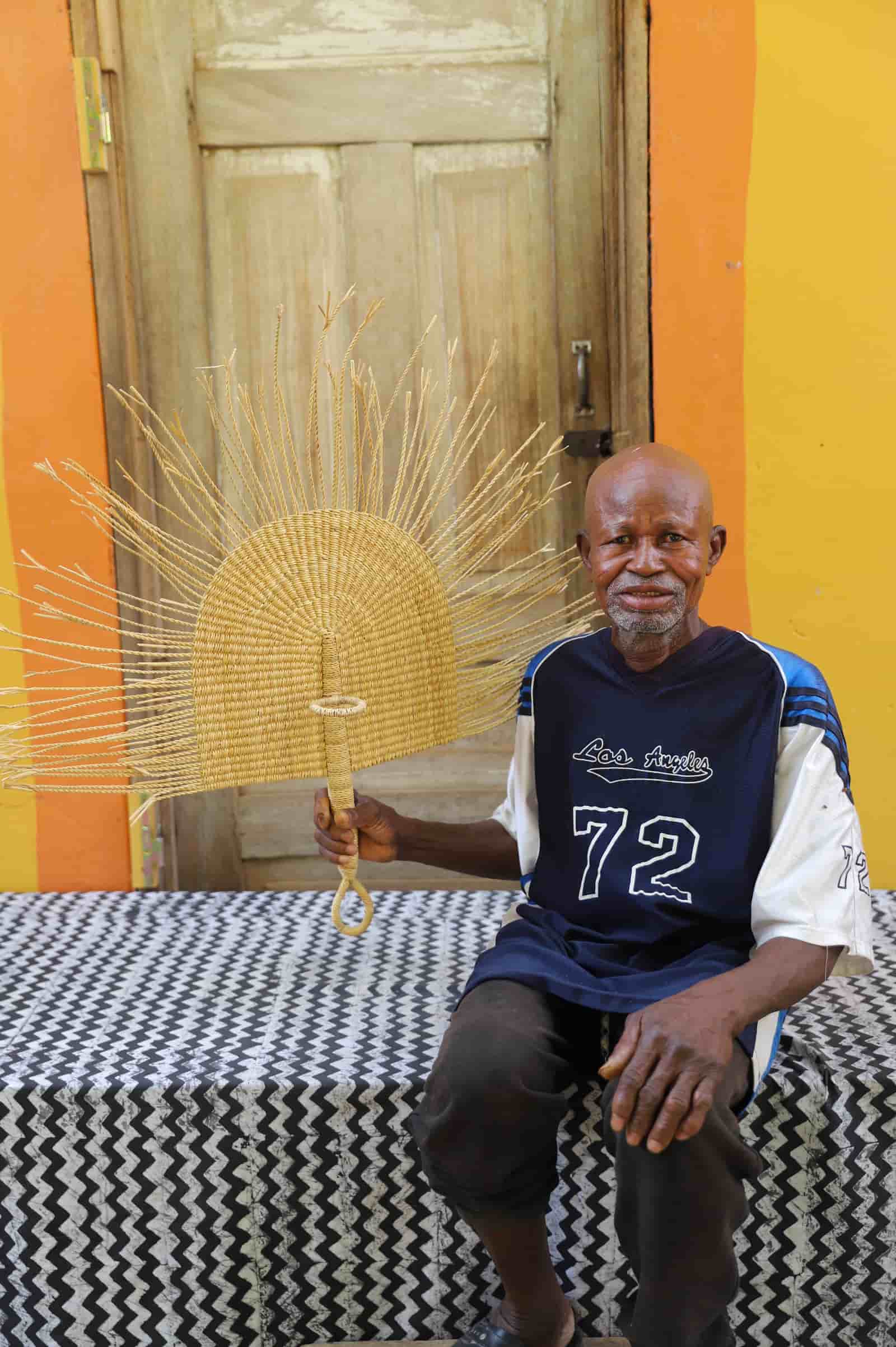 Man sitting on a bed holding a fan against a colorful wall.