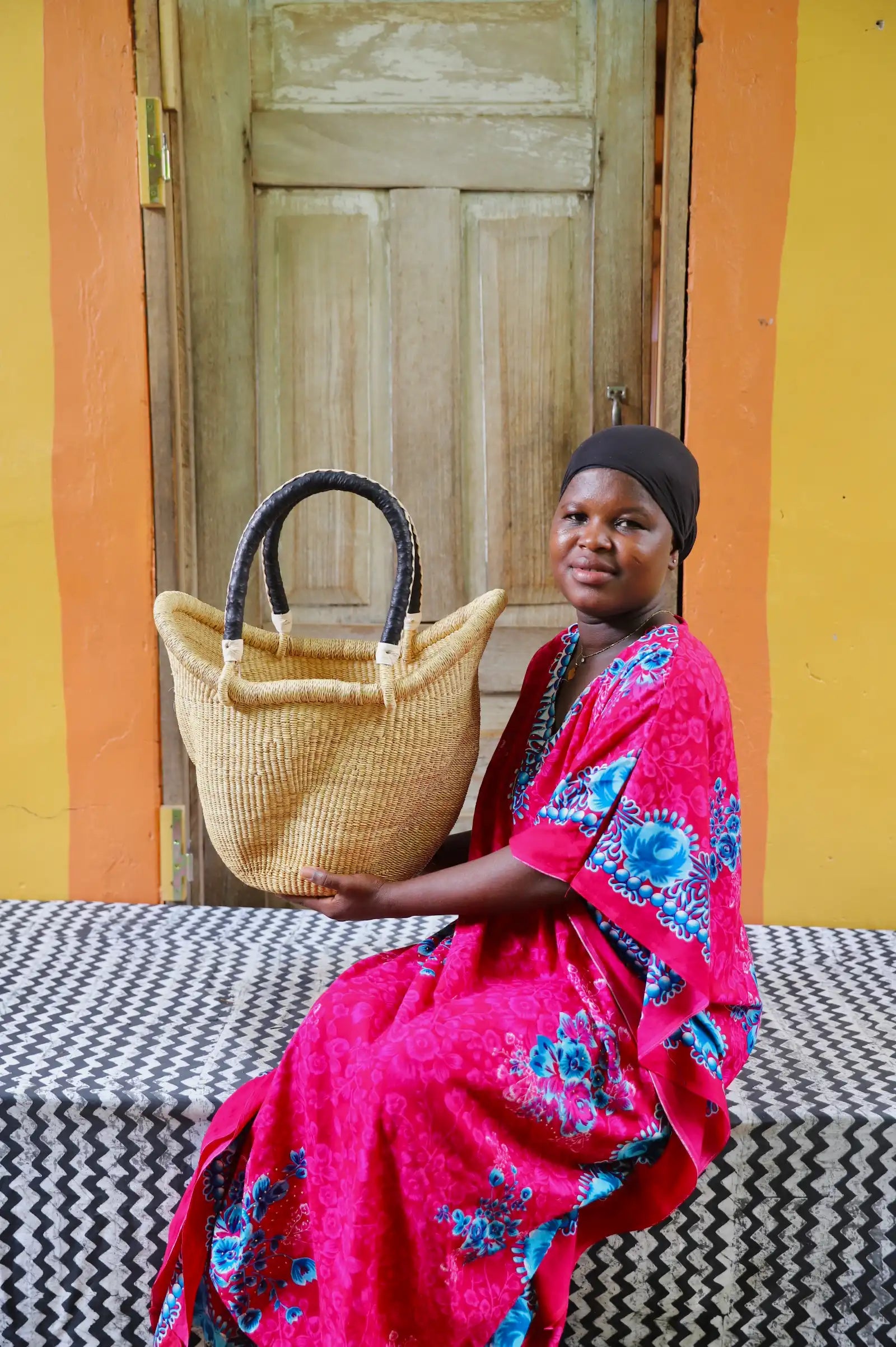 Woman in red floral dress seated holding a small natural Solara Carryall with dark leather handles
