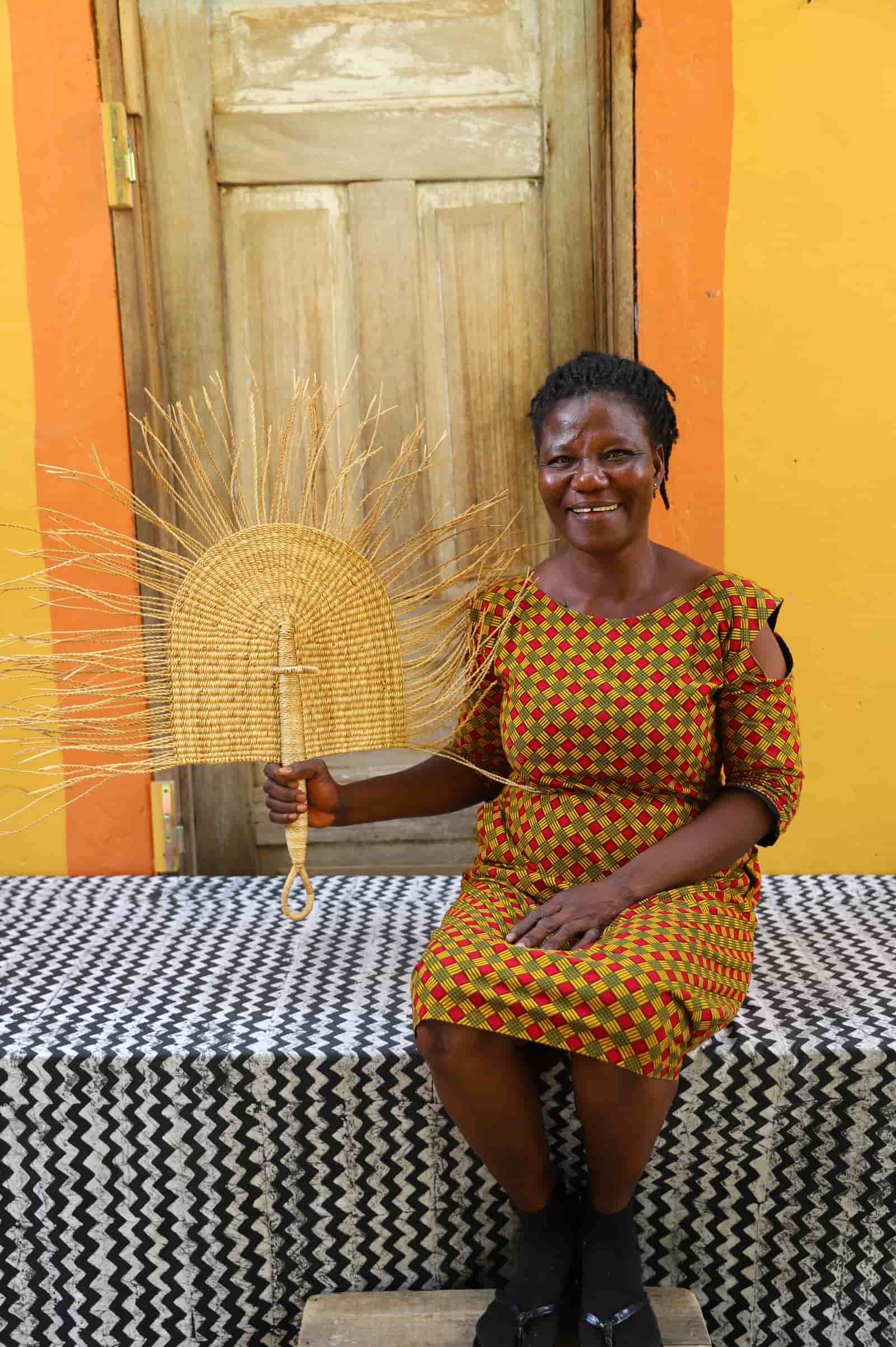 Woman holding a woven sun-shaped object against a colorful wall.