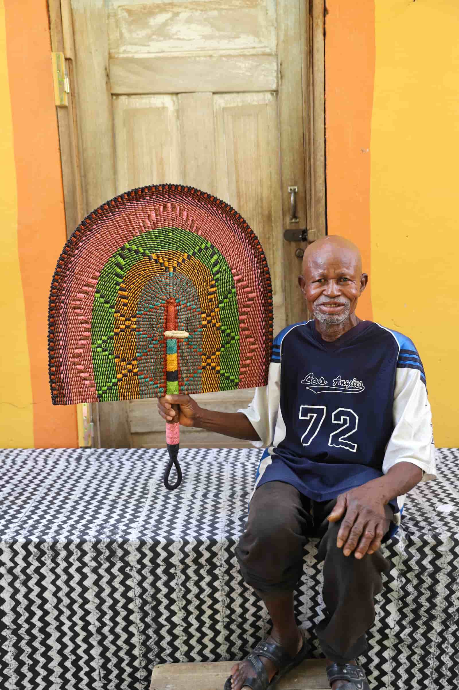 Man sitting on a black and white cloth  holding a colorful woven object against a yellow wall.