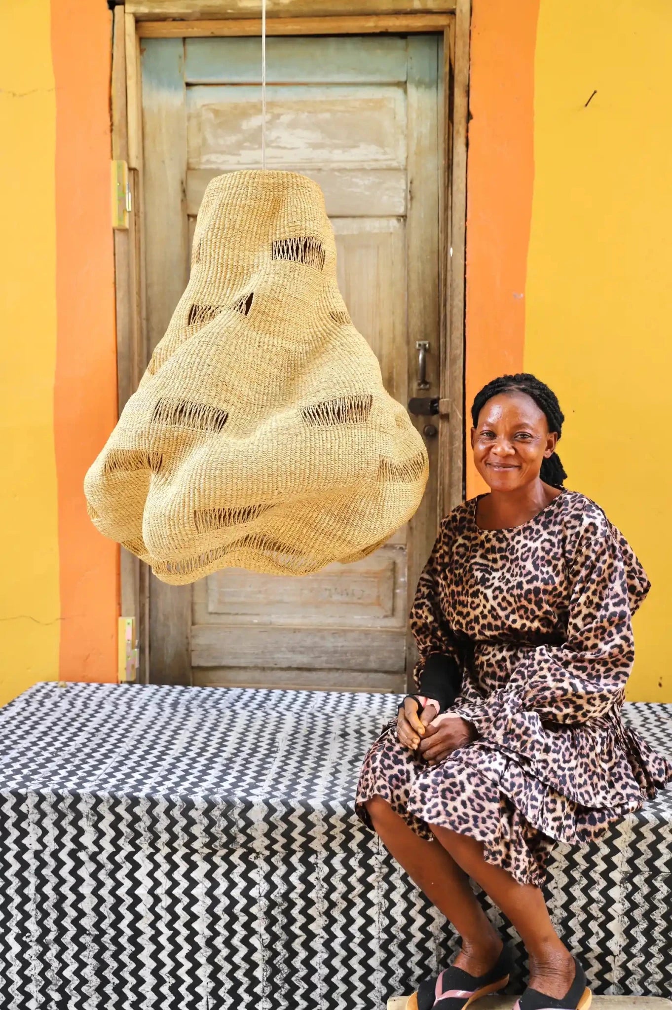 Woman seated beside a handwoven Cosma pendant lampshade in natural straw with an open weave, before a wooden door.