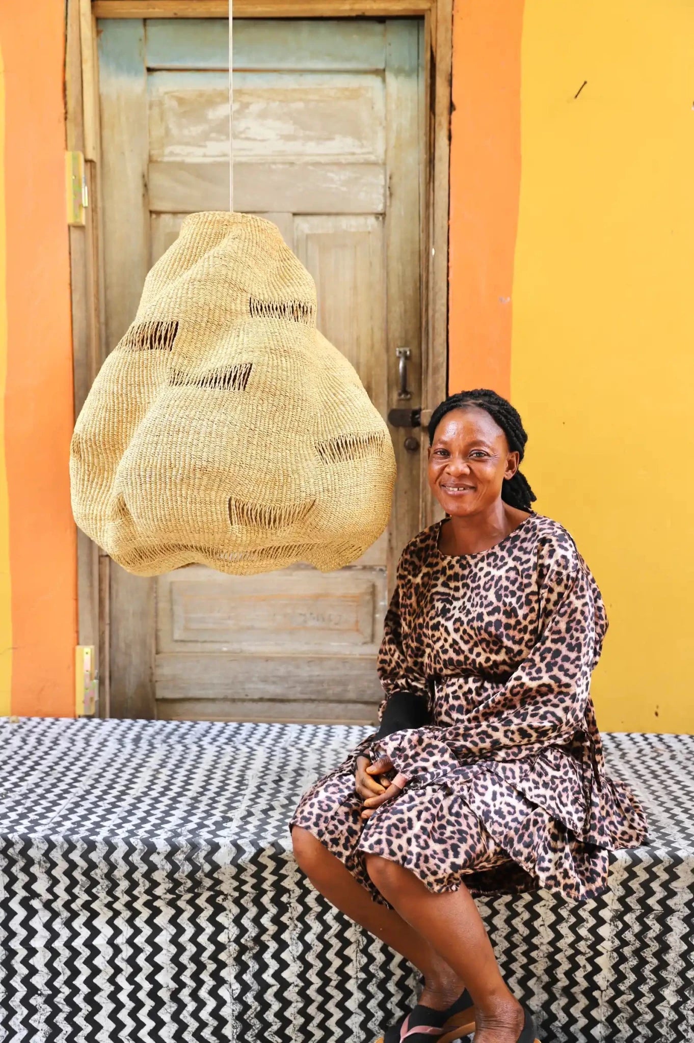 Woman in a leopard print dress sitting on a bench with a woven bag behind a wooden door.