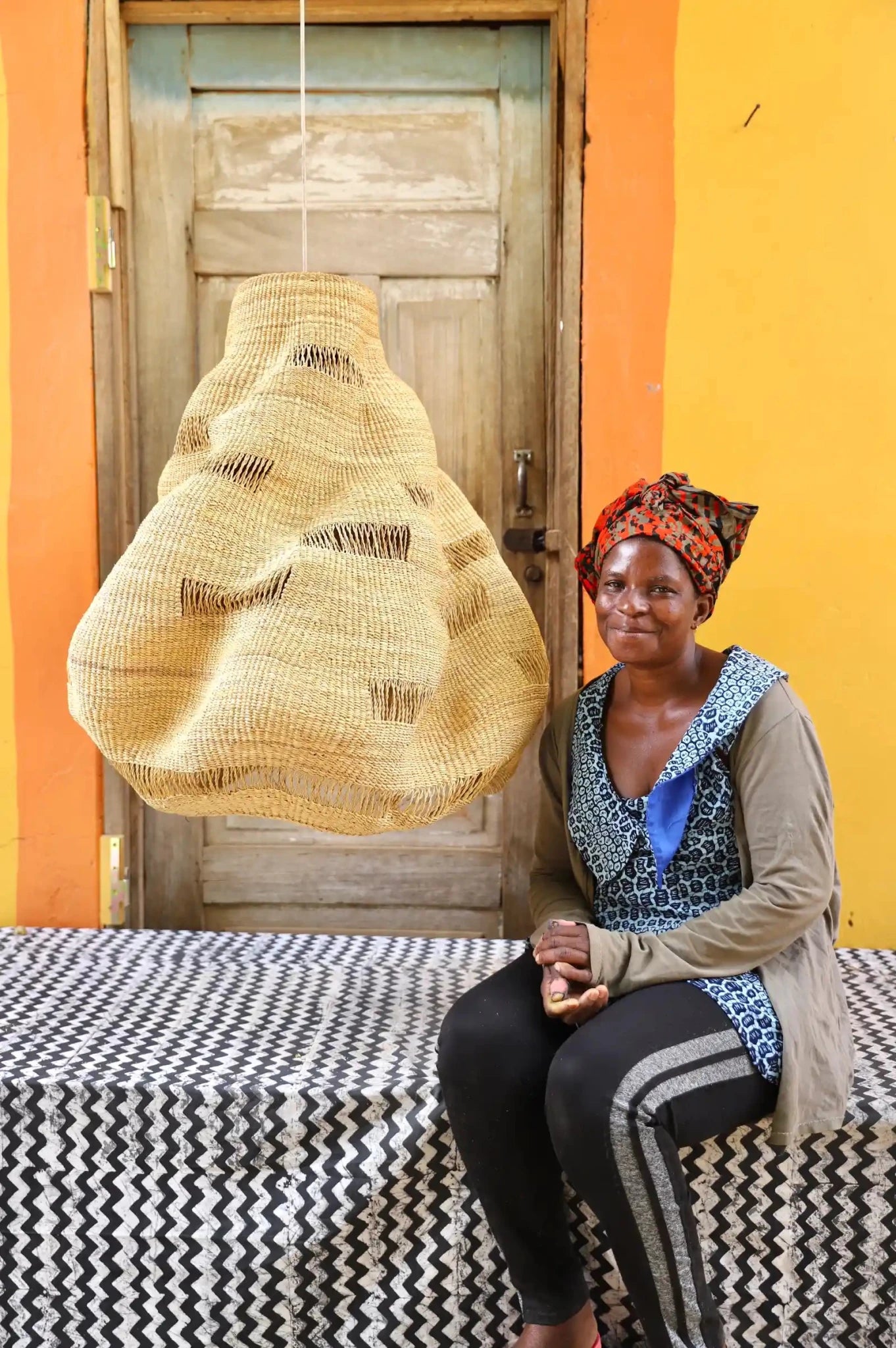 Woman sitting on a bed with a colorful headscarf and patterned fabric, against a vibrant yellow wall.