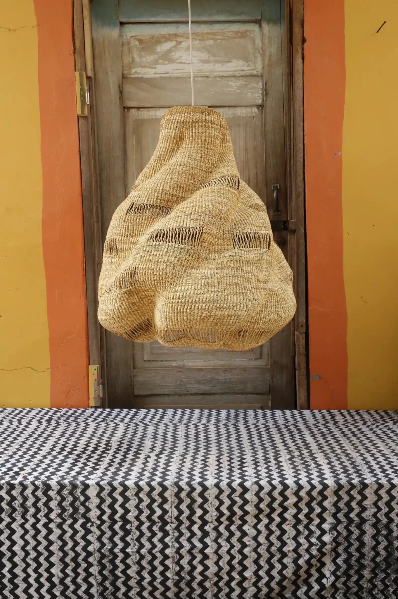 Woven basket hanging above a table with a patterned tablecloth against a yellow wall and wooden door.